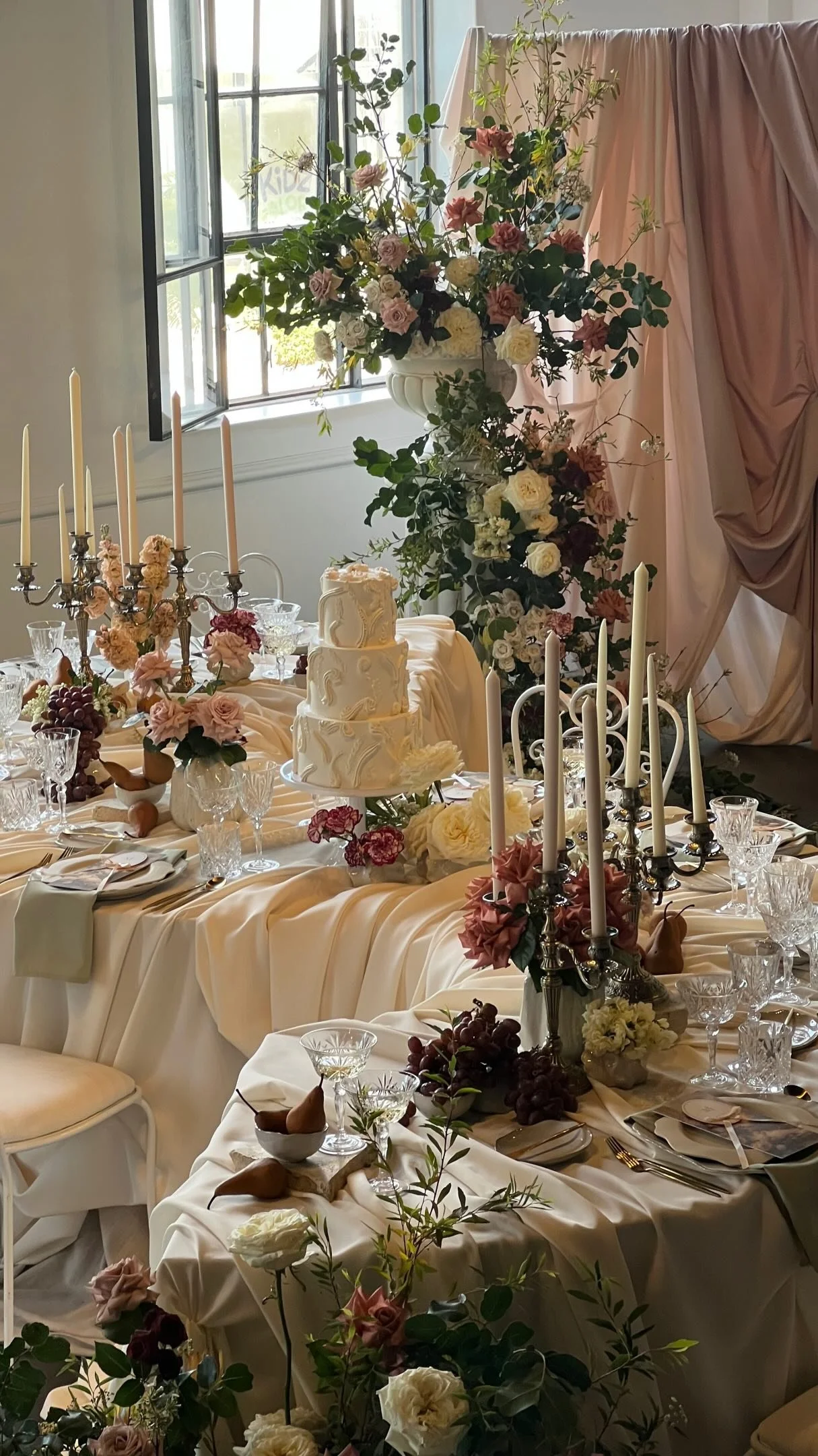 Elegant wedding reception table with a tiered wedding cake, surrounded by flowers and candle holders, near a window with curtains.
