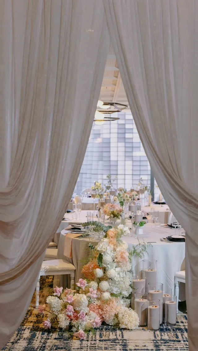 Beautiful banquet table with floral arrangements, candles, and place settings, seen through cream-coloured curtains in a modern, well-lit venue. Candle styling by Candlescapes, Perth Candle Styling & Hire.