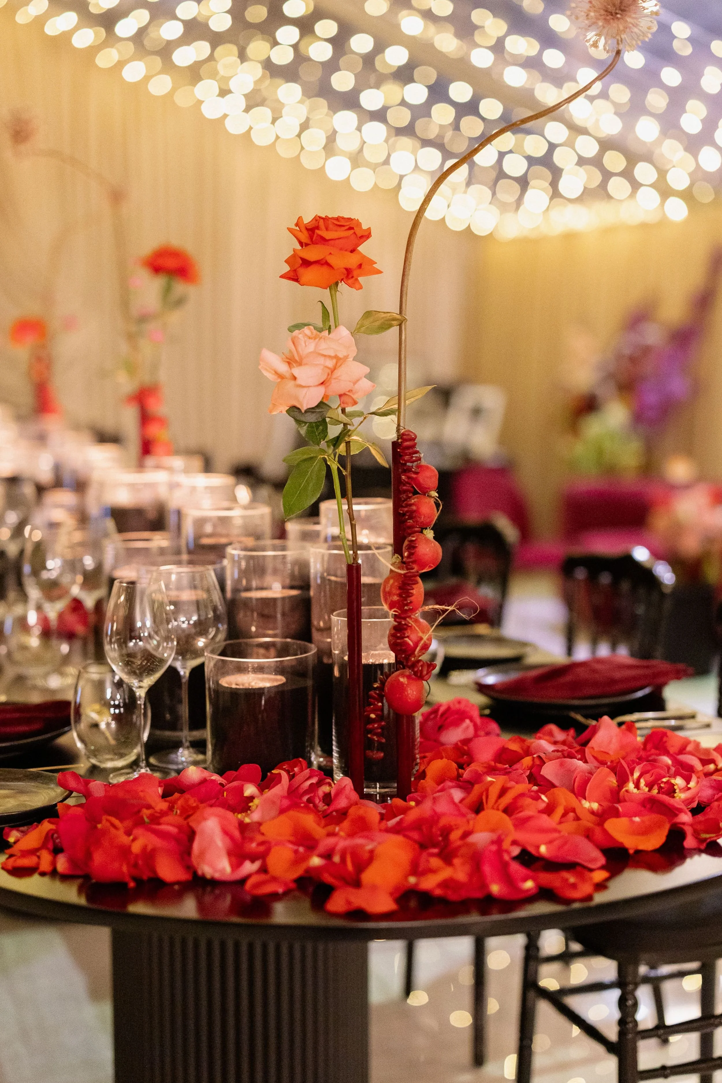 Photo by Tessa Kit Photography. A floral centrepiece featuring orange and pink roses with rose petals scattered on a table, set for a celebration or formal event in a decorated venue with blurred tables and fairy lights in the background.
