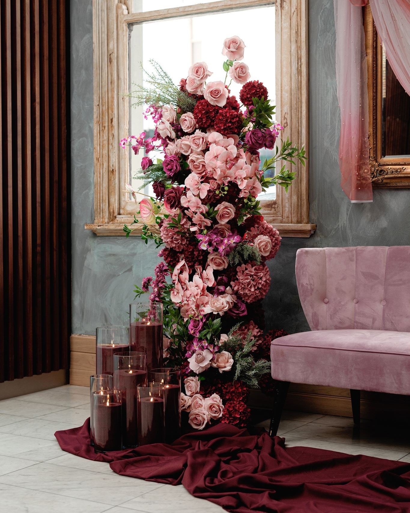 A large pink and red floral arrangement with roses, hydrangeas, and other flowers is placed inside a room by a window. There are several tall candles in glass holders on the floor, surrounded by a burgundy cloth. A pink upholstered chair is next to the arrangement, and the room has a gray wall with a gold-framed mirror and pink drapes.