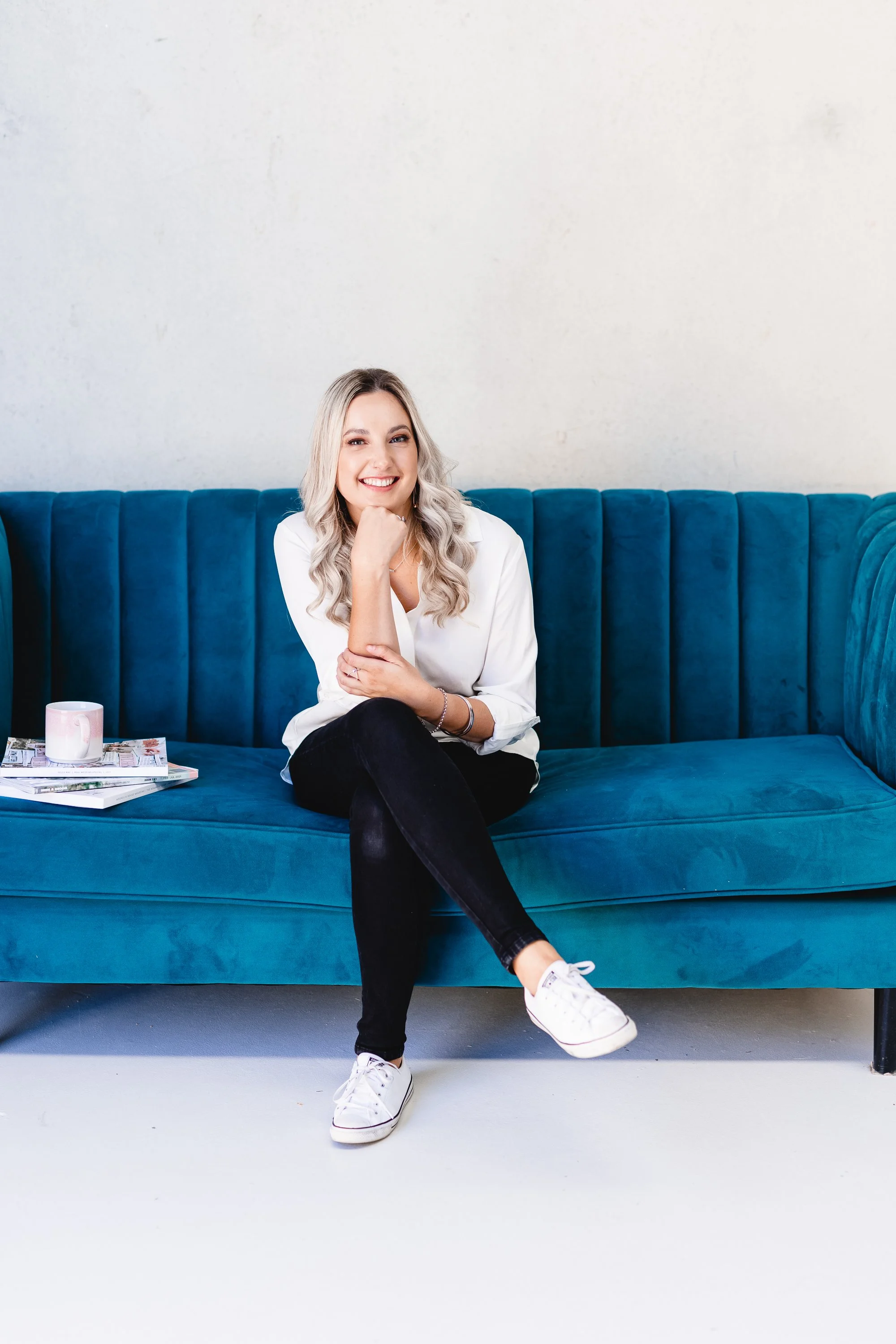 Aimee Shadbolt, of Candlescapes, Perth.  She has long blonde hair and is smiling and sitting on a blue velvet sofa, wearing a white top, black pants, and white sneakers, with magazines and a mug on the sofa beside her.