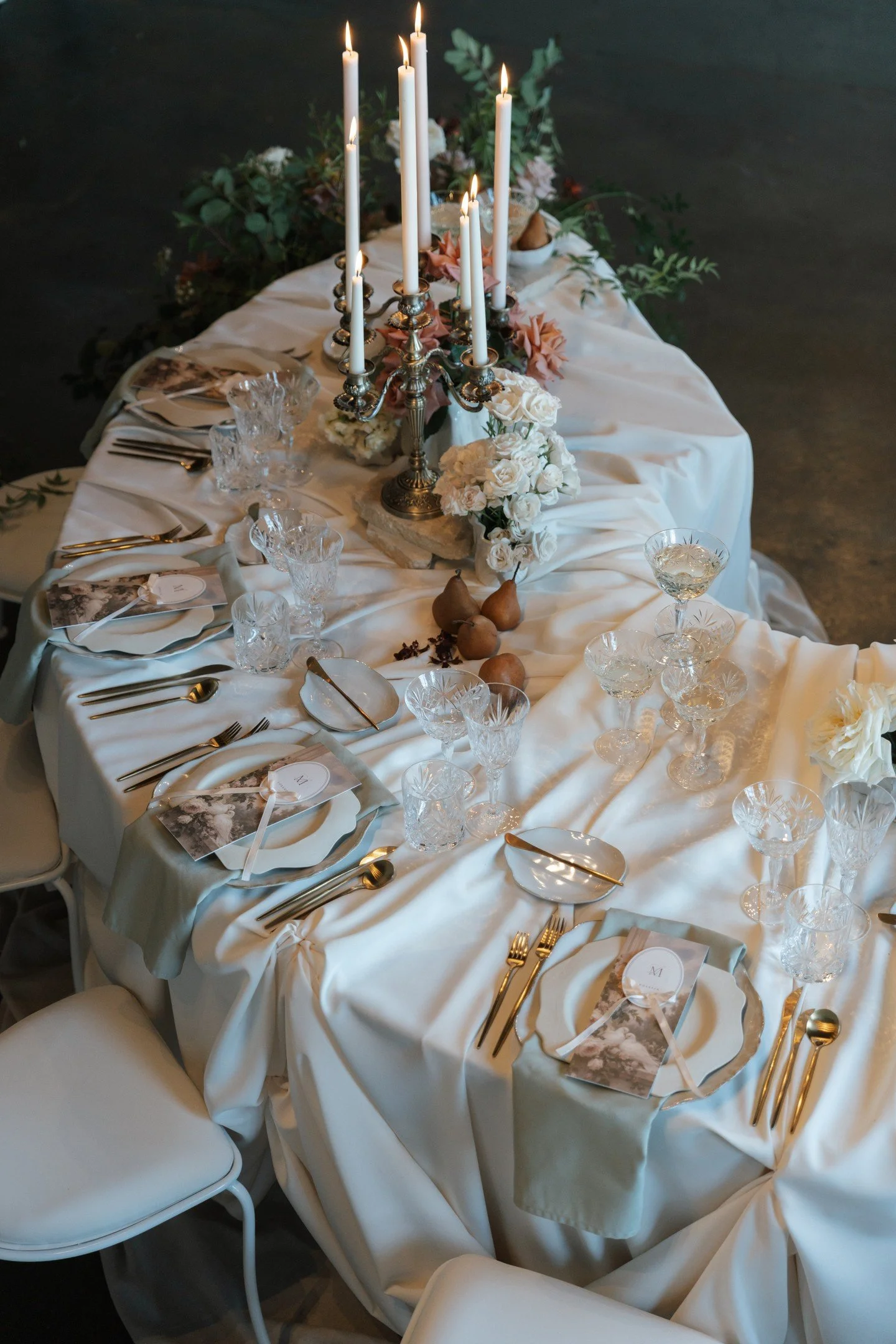 Elegant rectangular dining table draped with white cloth, set with crystal glassware, gold utensils, and printed place cards, with a vintage candelabrum holding white candles and floral arrangements, in a dimly lit room.