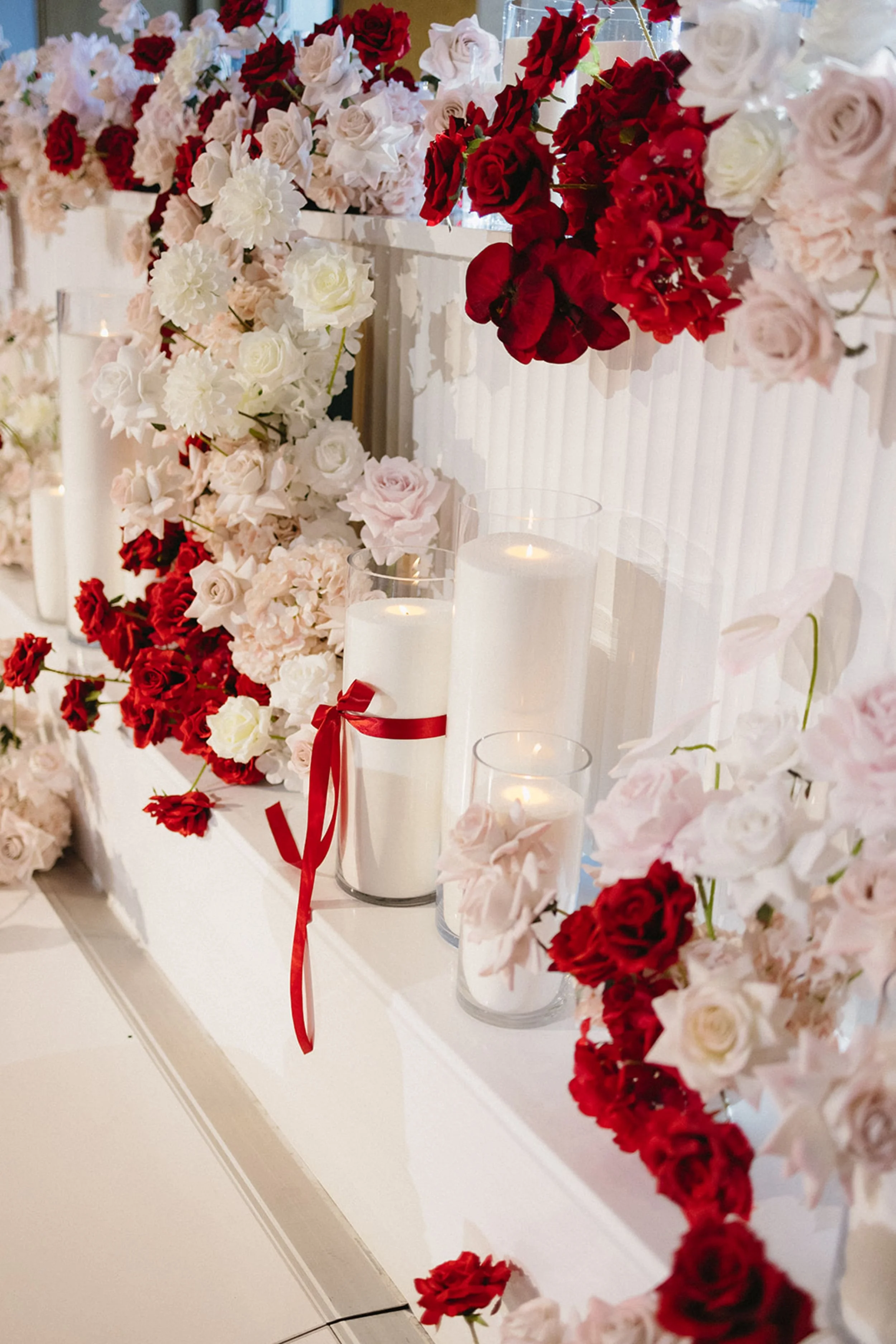 Photo by Mitch & Tijana.  Flowers in shades of red, pink, and white arranged along a white surface, with white candles in glass holders tied with red ribbons among the flowers.