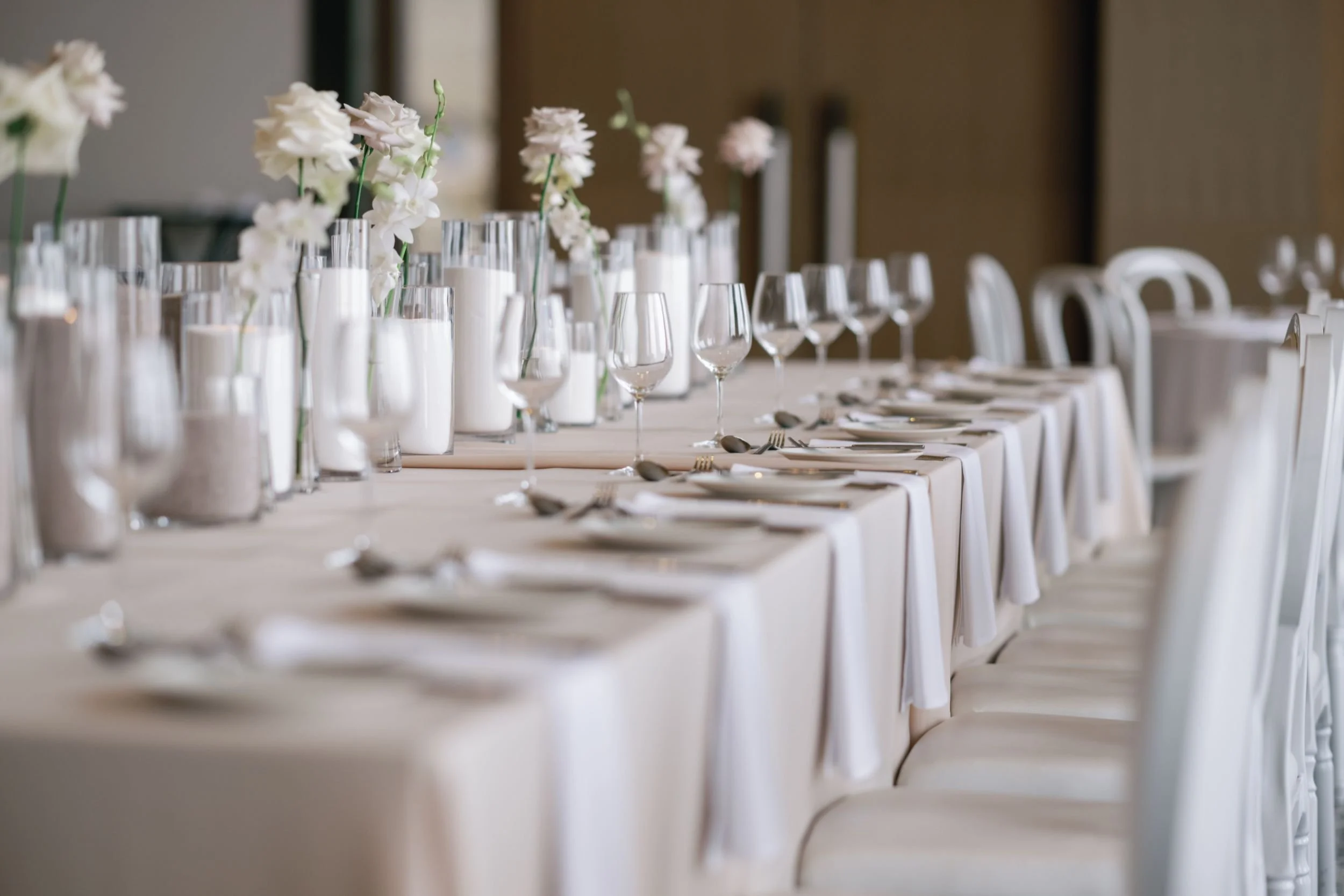 Photo by Norman Yapp Photography.  Wedding reception table with white tablecloths, decorated with white flowers in glass vases, and set with silverware and wine glasses.