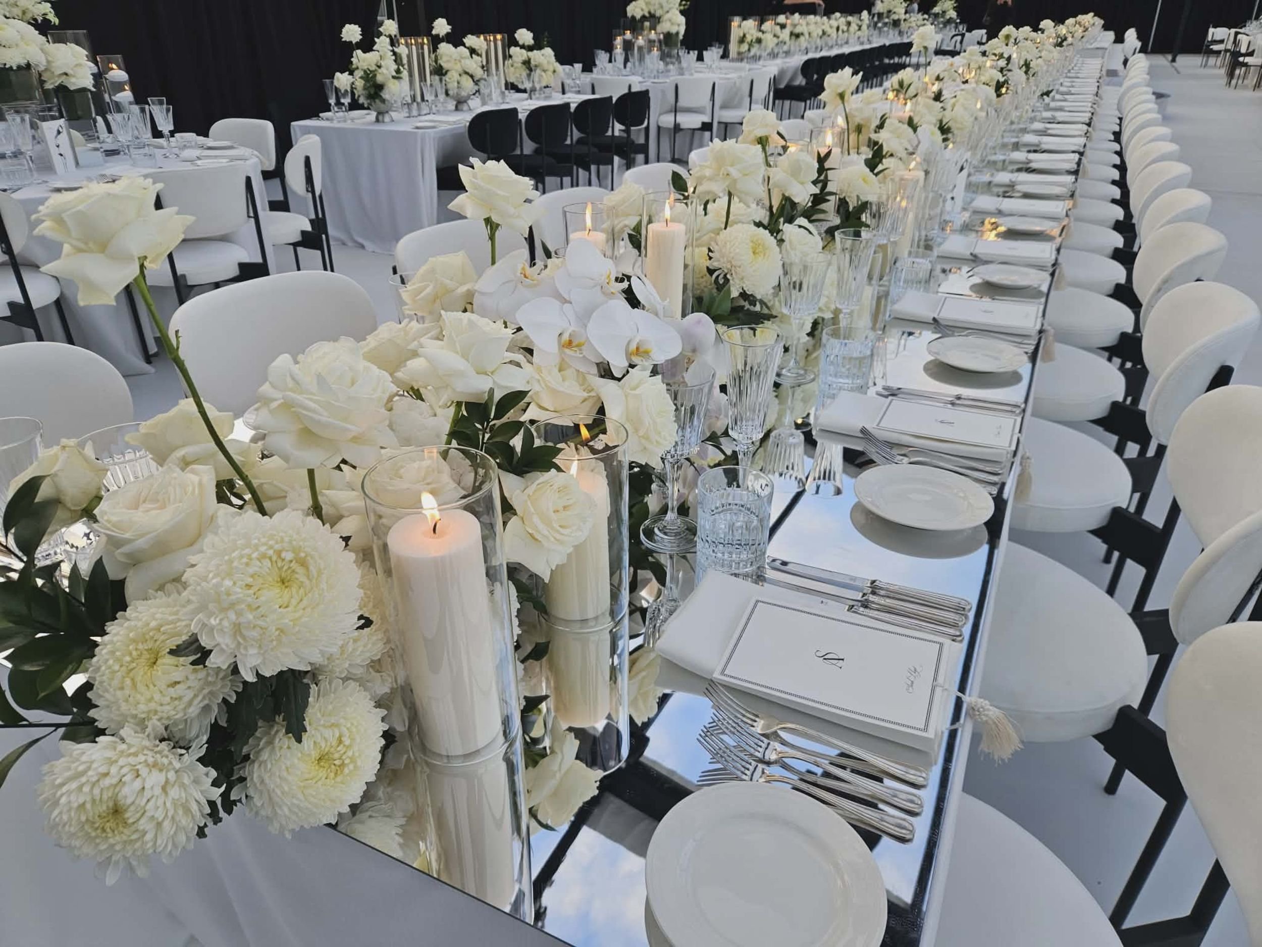 Wedding reception table decorated with white flowers, candles, and glassware, with white chairs arranged along the length of the table.