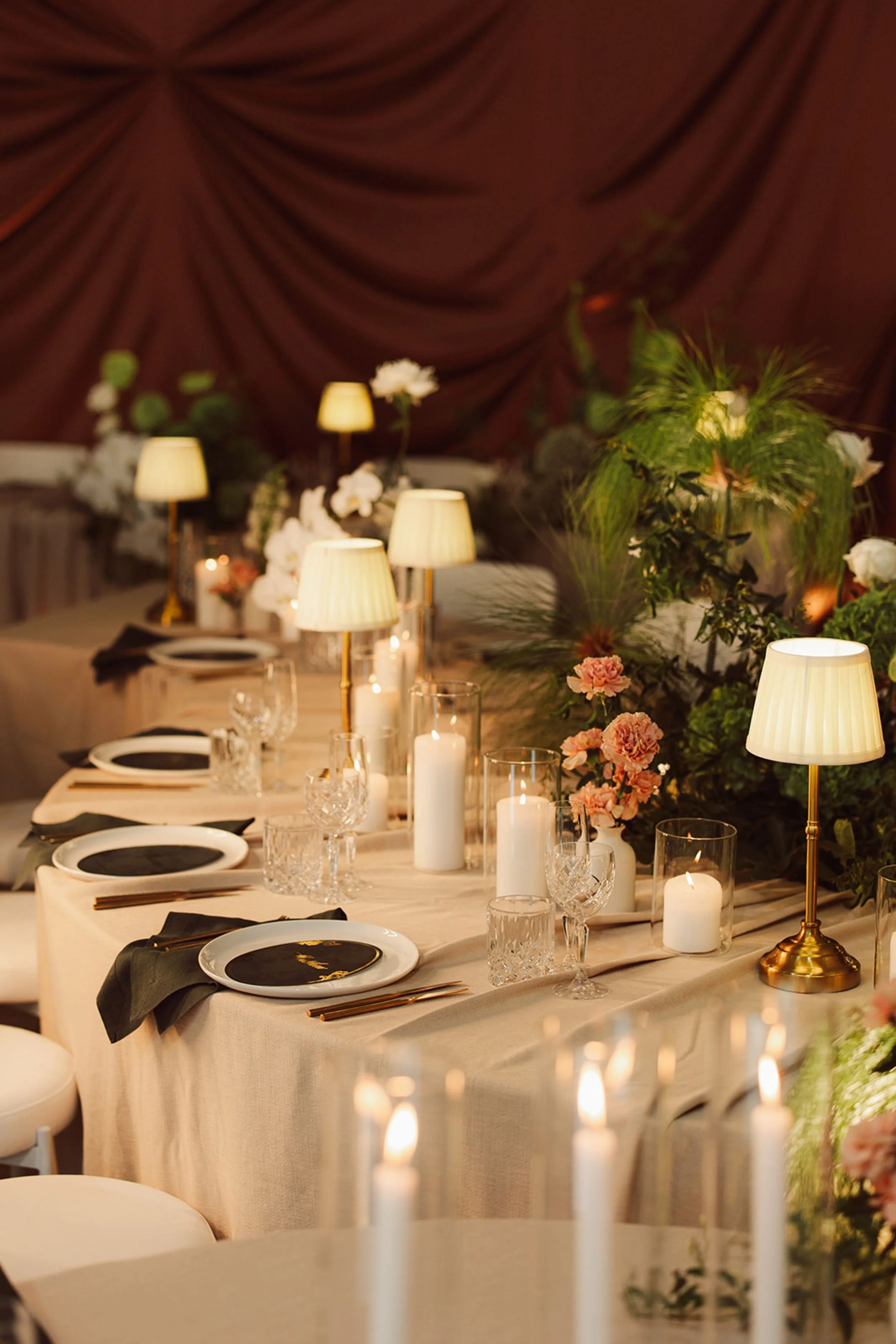 Photo by Jamie Lee Photography.  Elegant dining table decorated with flowers, candles, and tableware, set for a formal event in a room with draped maroon curtains and soft lighting.  Candle styling by Candlescapes, Perth Candle Styling & Hire.