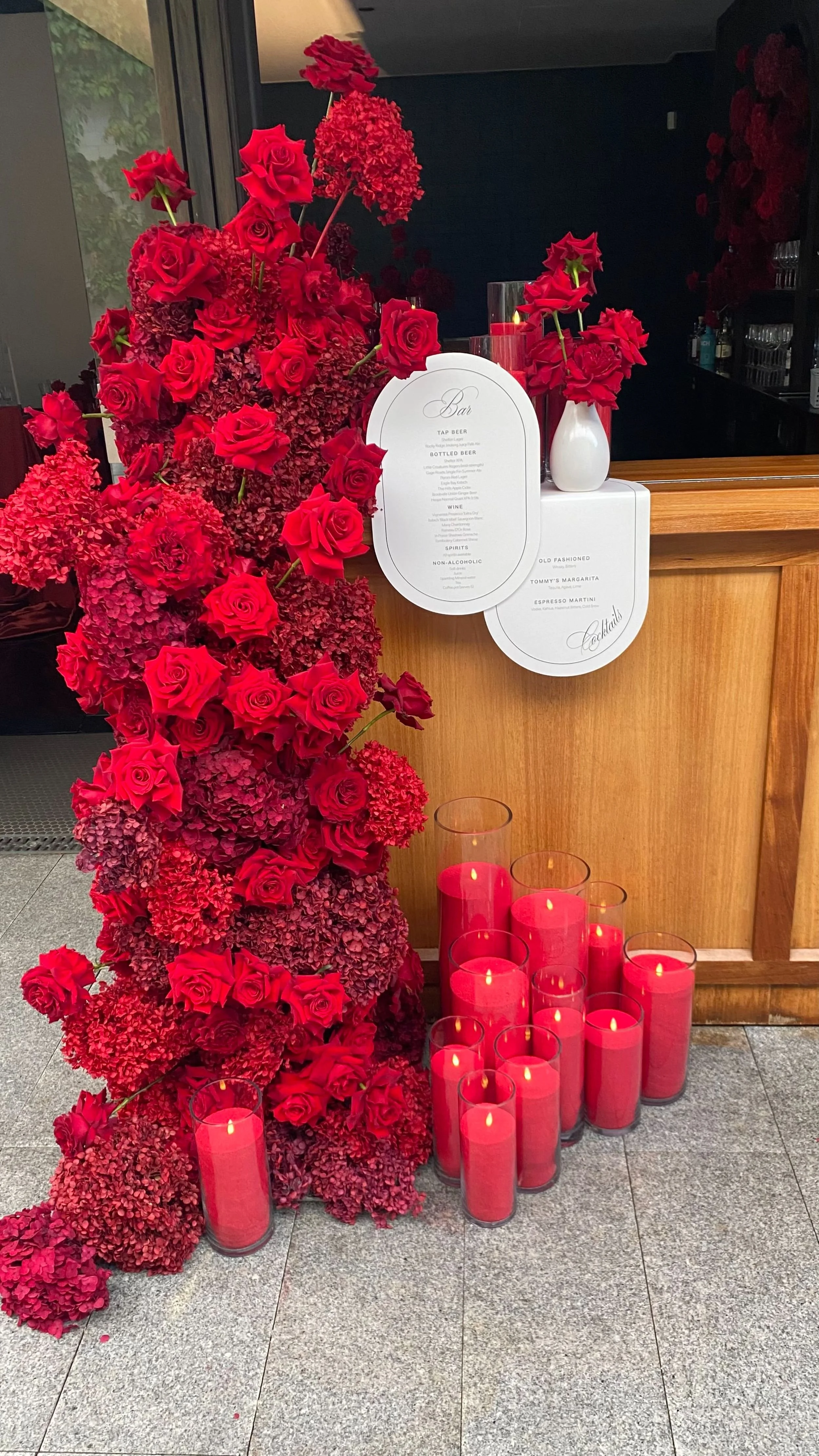 A floral display with red roses and hydrangeas arranged on a stand, accompanied by red candles in glass holders and two vases with additional red flowers. A menu is hung on a wooden counter in the background.