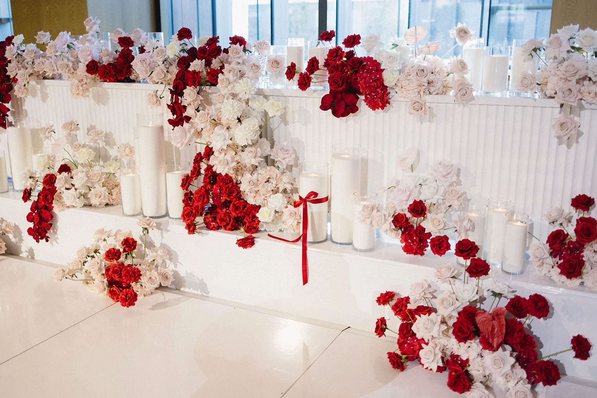 Photo by Mitch & Tijana.  A decorative arrangement of red and white flowers on a table, adorned with candles in glass holders and some with red ribbons, set against a backdrop of large windows.