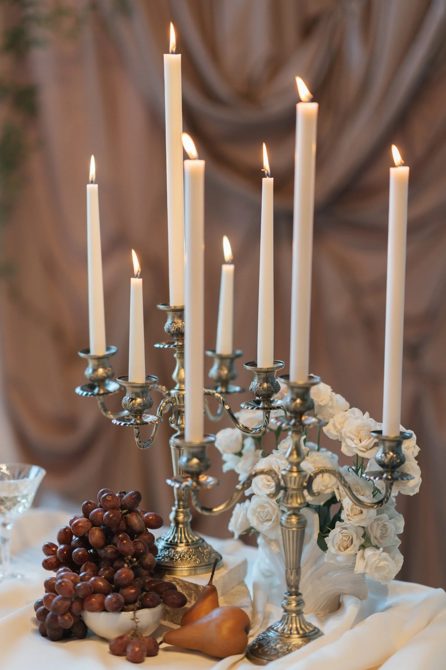 A decorative table setting with a silver candelabra holding seven lit white candles, surrounded by white roses in a vase, with grapes and pears on a white cloth.