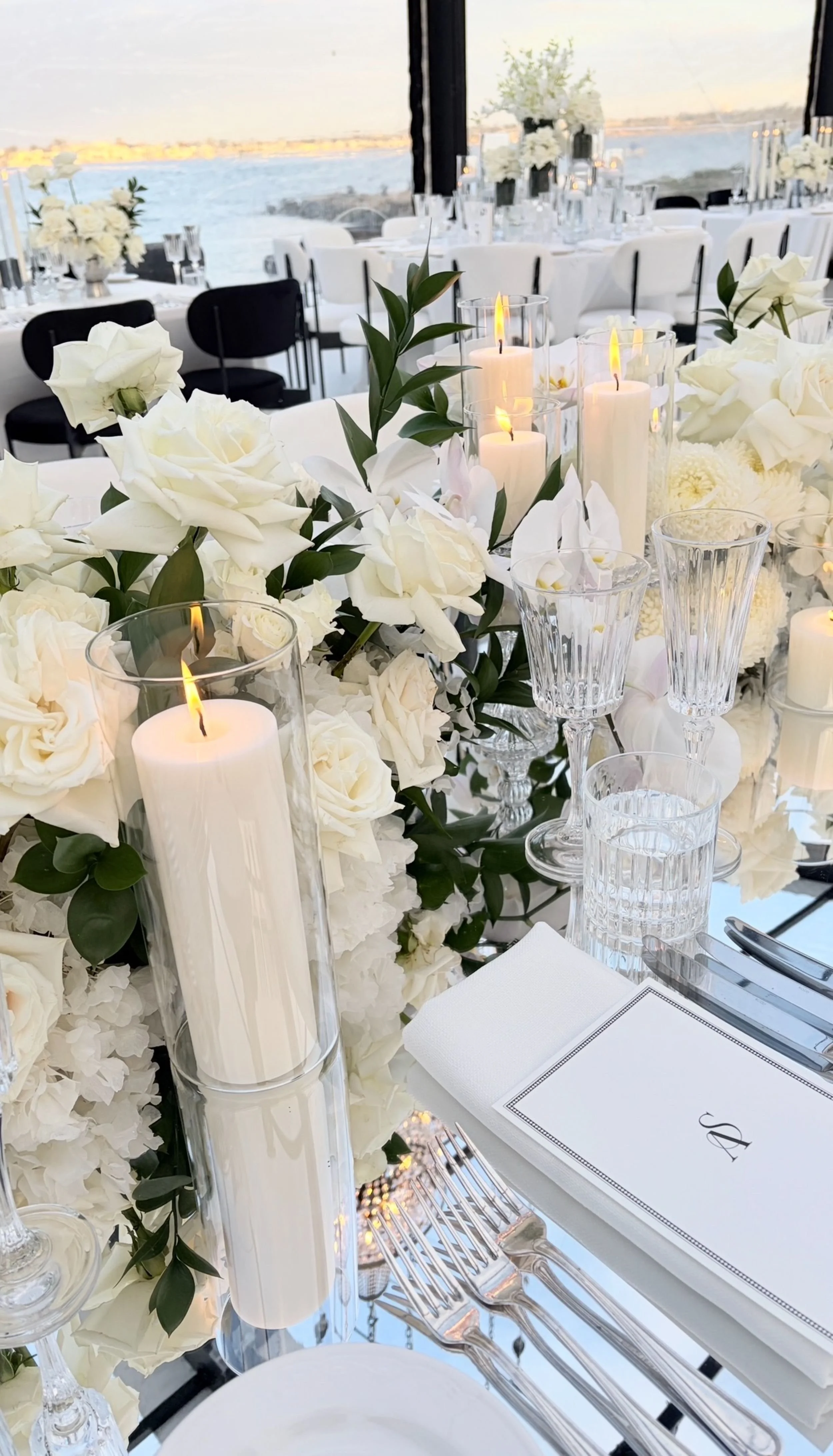 Elegant wedding or formal event table setting with white roses, lilies, and greenery, tall white candles in glass holders, crystal glassware, and a waterfront view in the background.