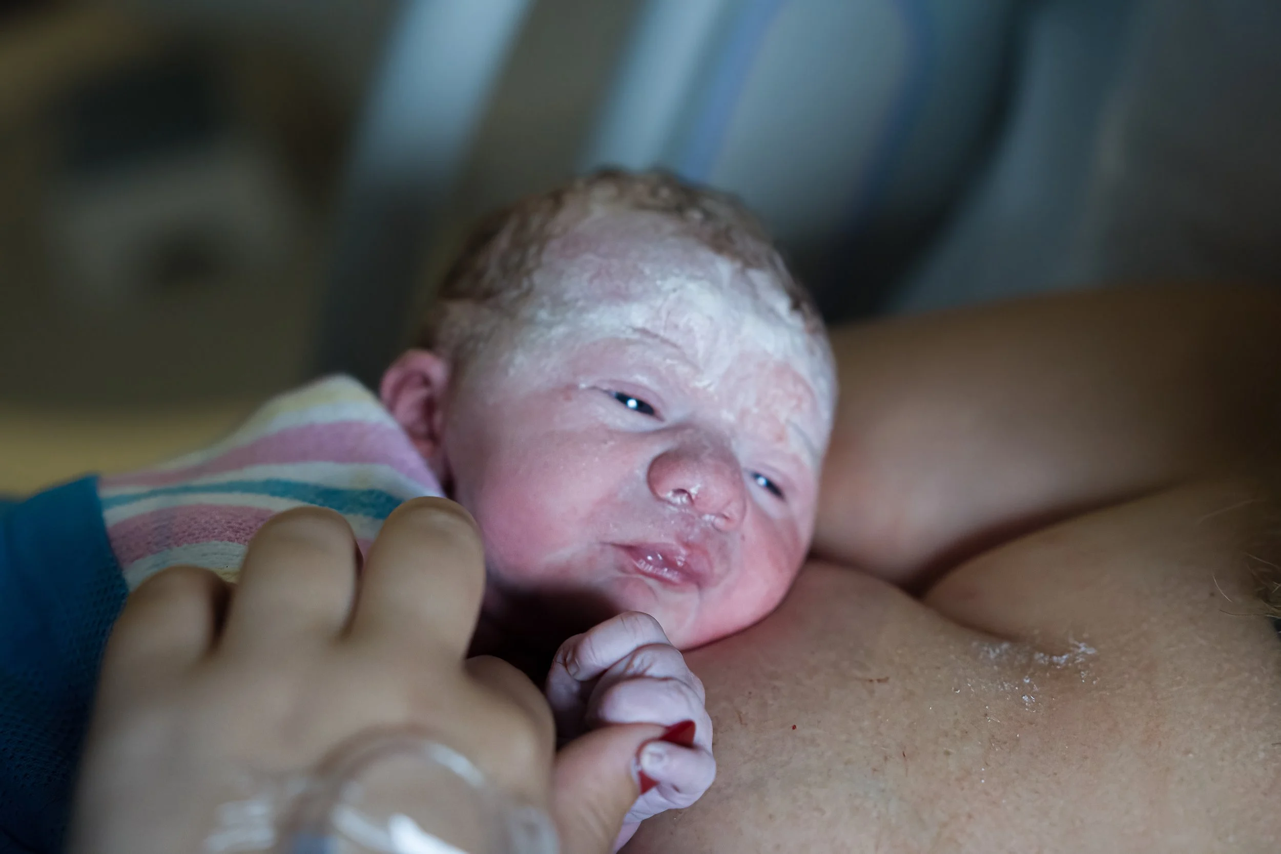 Newborn baby covered in vernix, being held and swaddled in a blanket, with a hospital setting background.