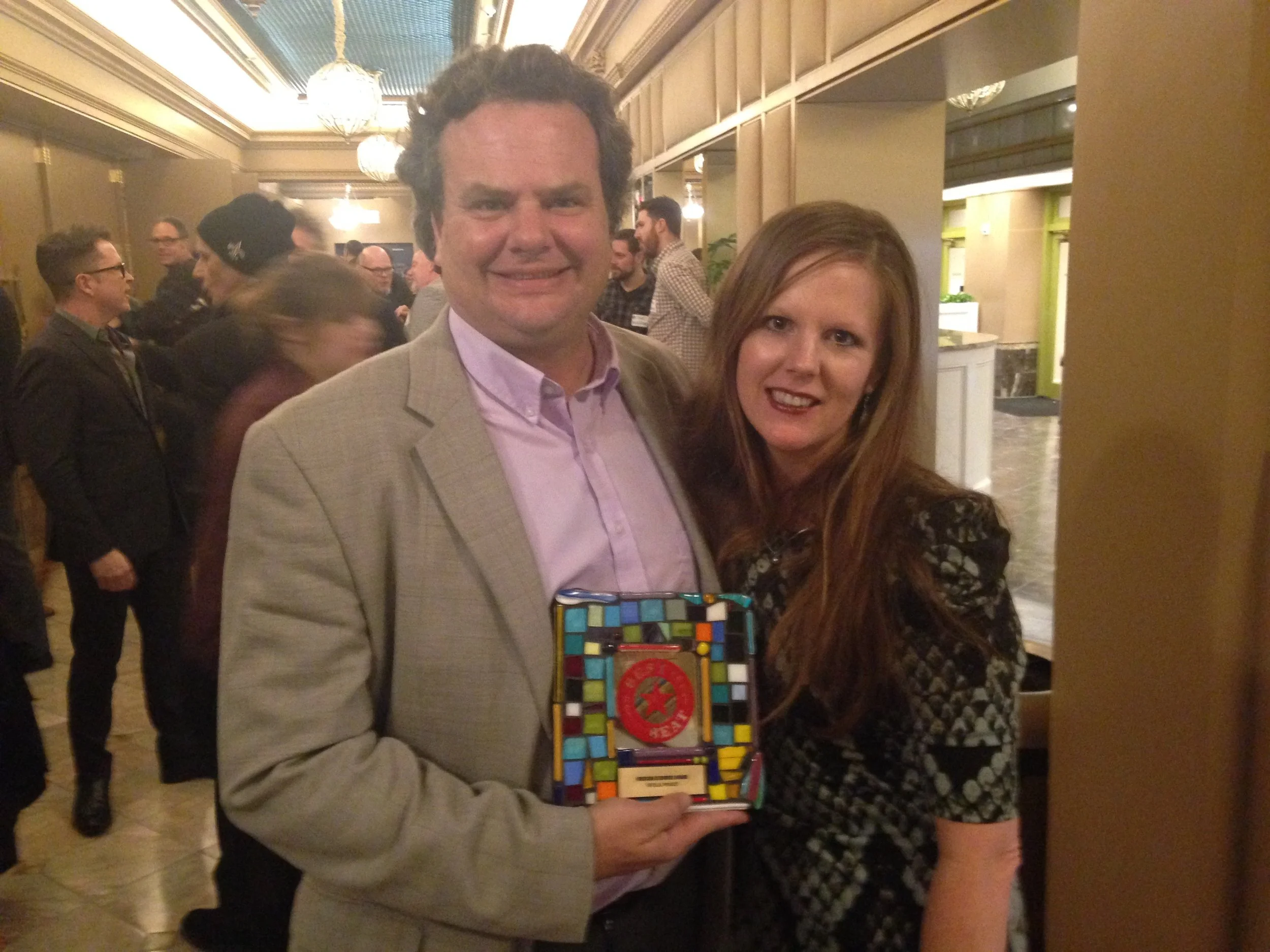 A man and woman at an event, holding a colorful award or plaque with a red star emblem, smiling at the camera.