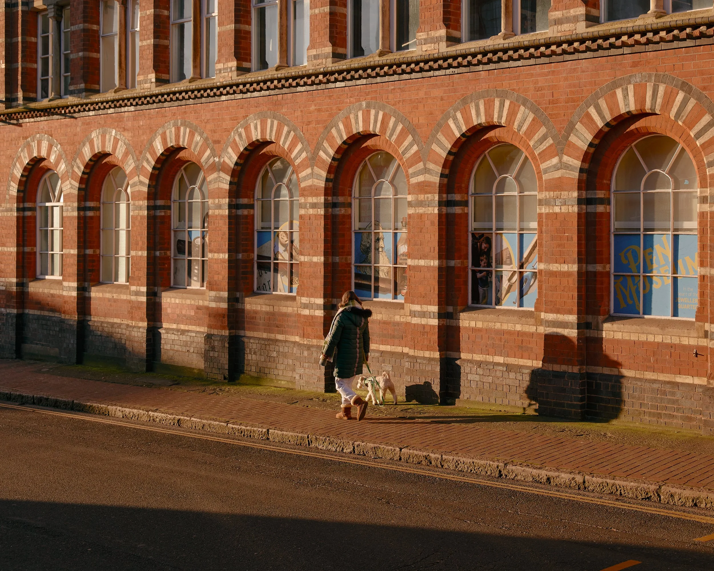 A woman walking her dog past red brick buildings in warm low light in the Jewellery Quarter, Birmingham.
