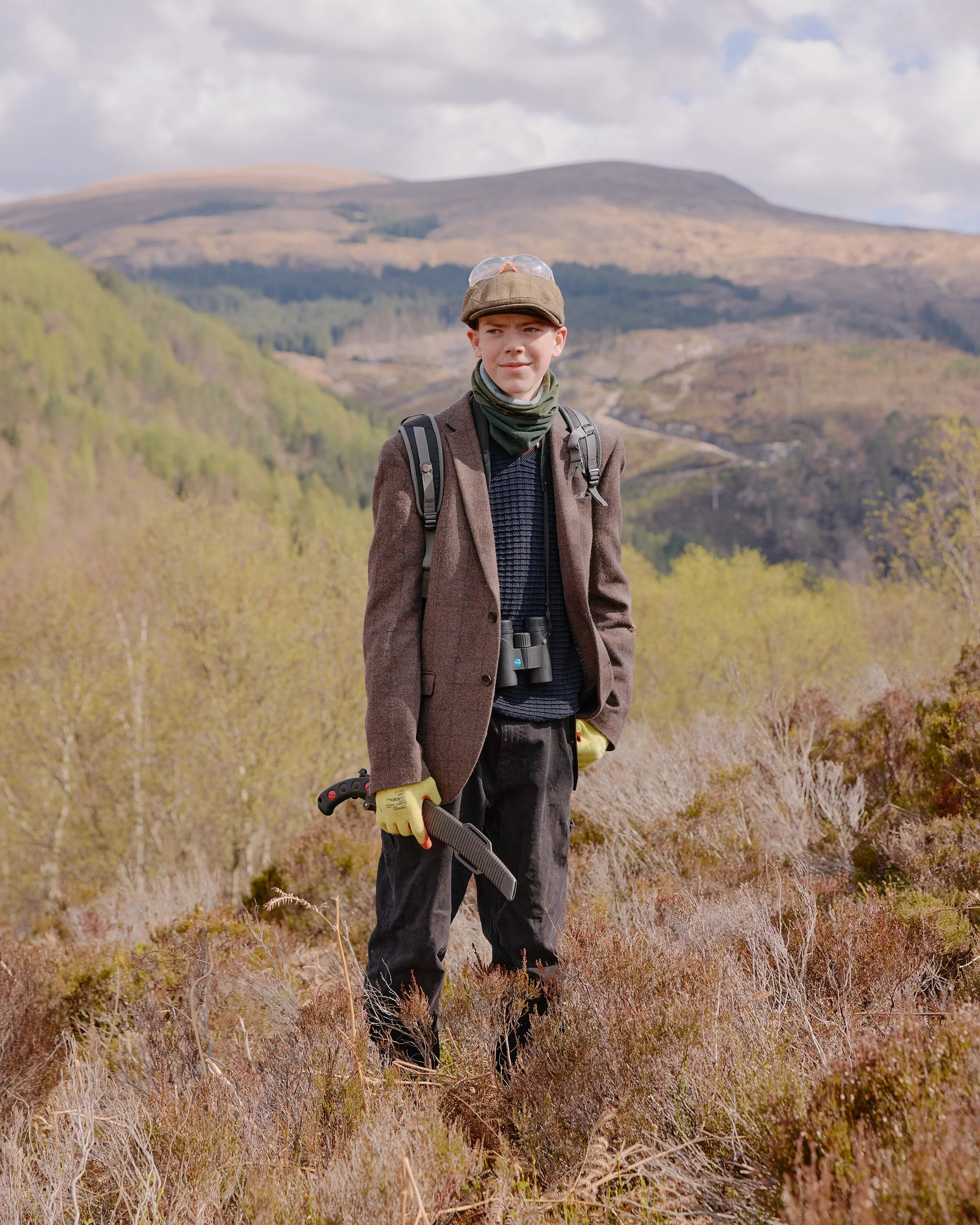 A teenager wearing a tweed jacket and flat cap stands on a hillside in the highlands, Scotland