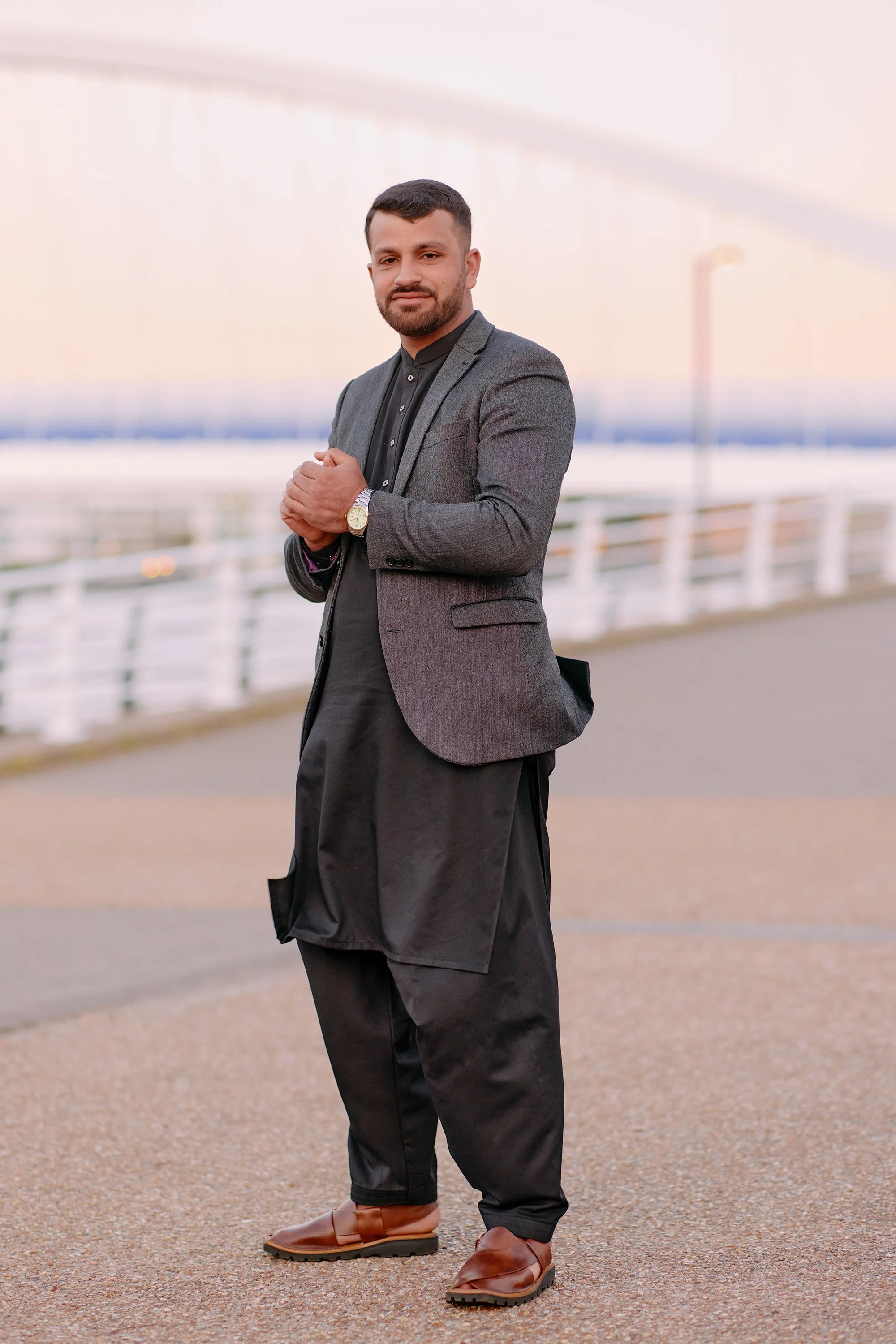 A man standing with his hands held together, looking toward the camera in soft evening light in Salford.