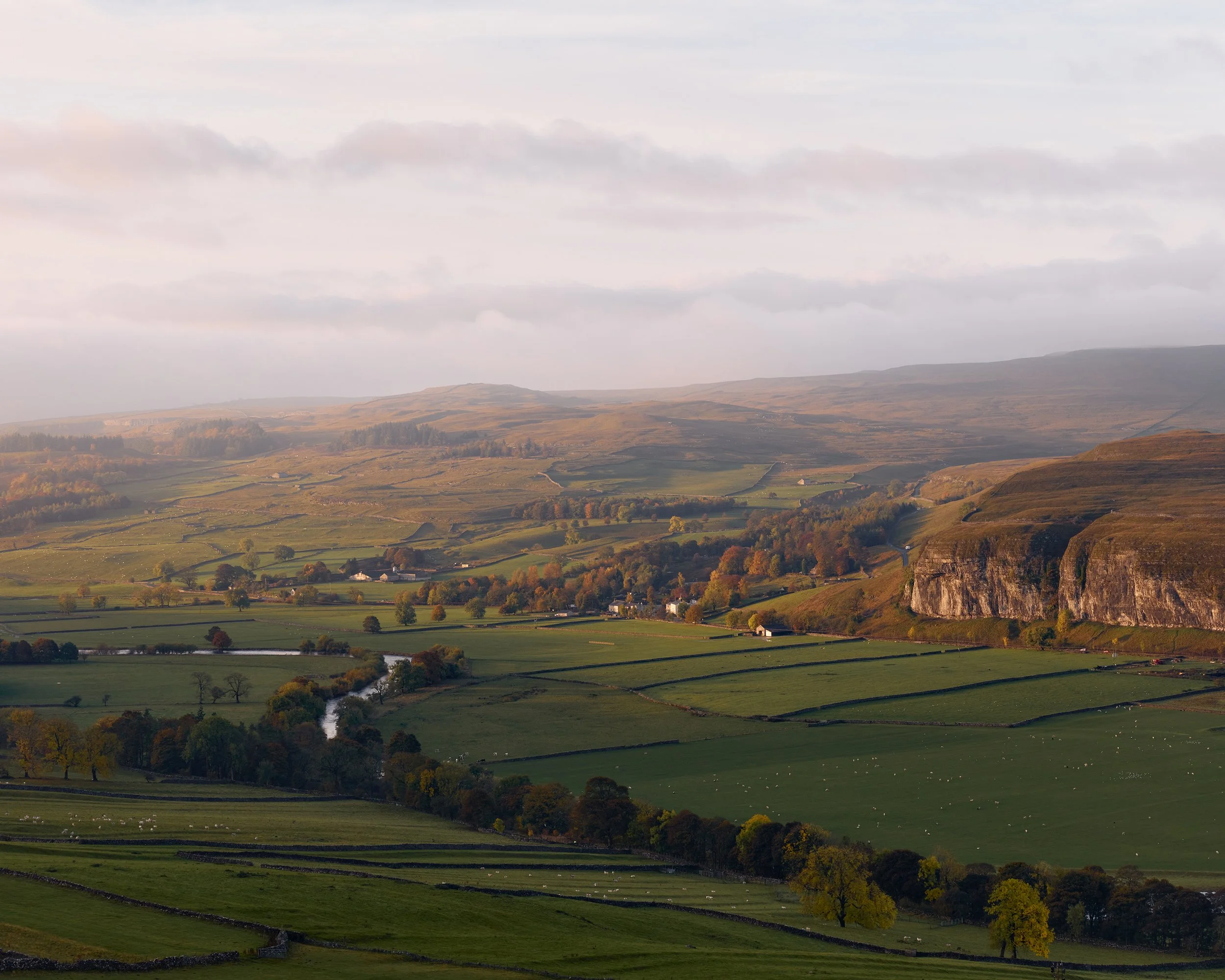 Warm morning light highlighting trees, buildings and the rocky cliff face of Kilnsey Crags.