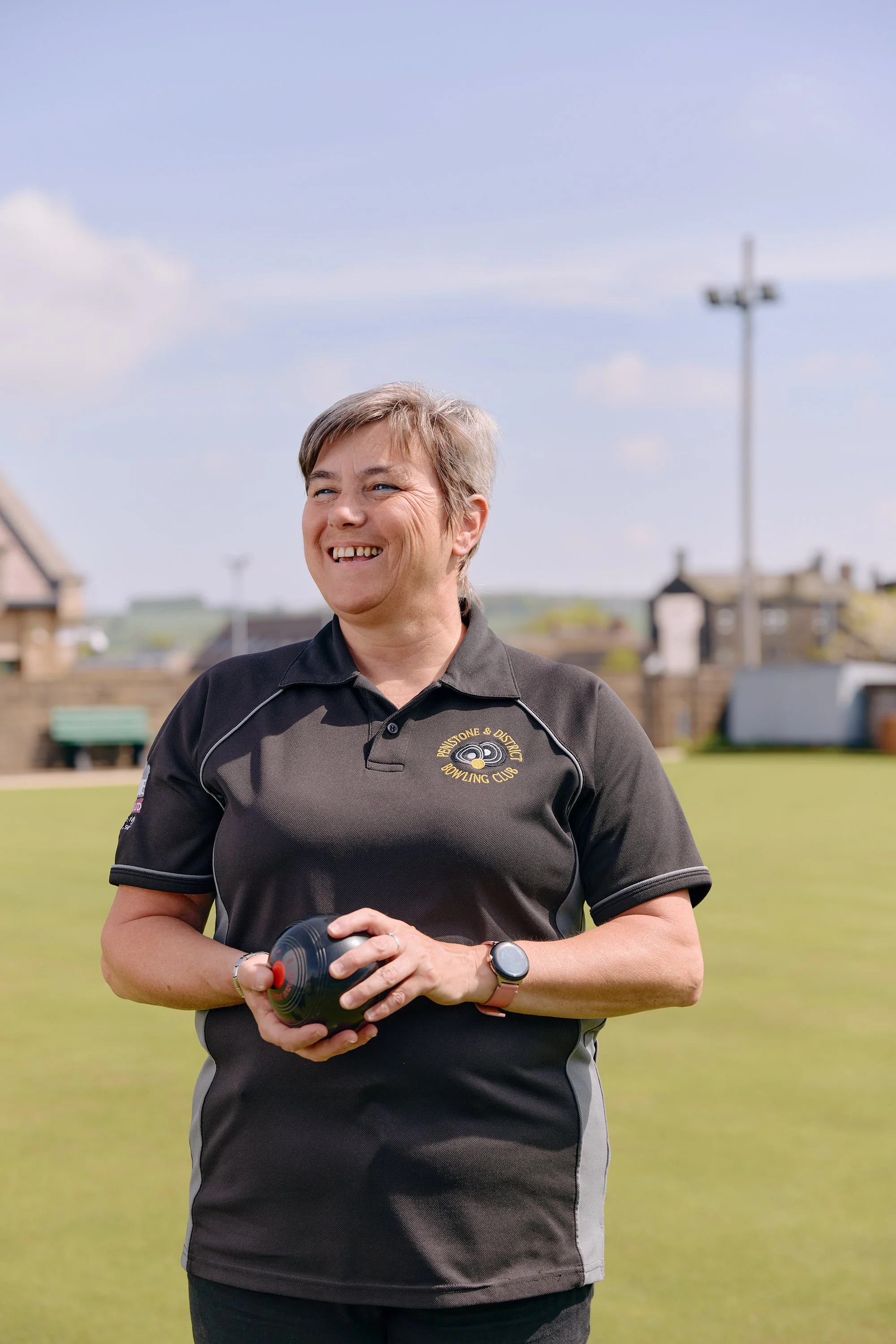 A woman smiling while holding a lawn bowl beside a bowling green in Penistone, England.