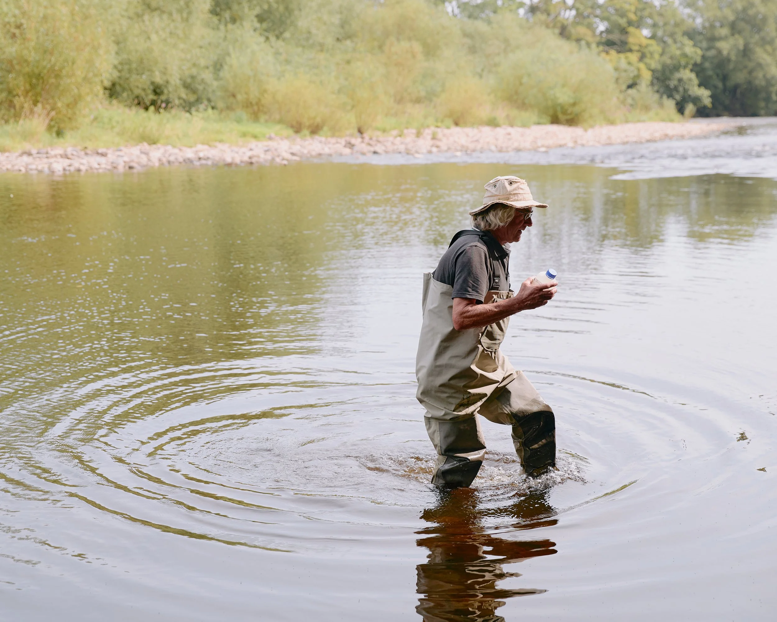 A citizen scientist wearing waders holding a water sample bottle while standing in the River Wharfe.