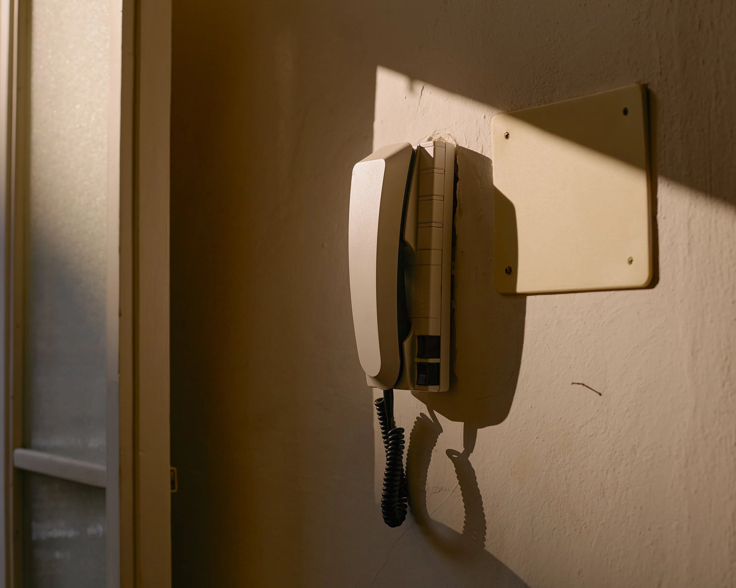 A wall-mounted corded telephone lit by angular sunlight and long shadows inside an apartment in Pisa.