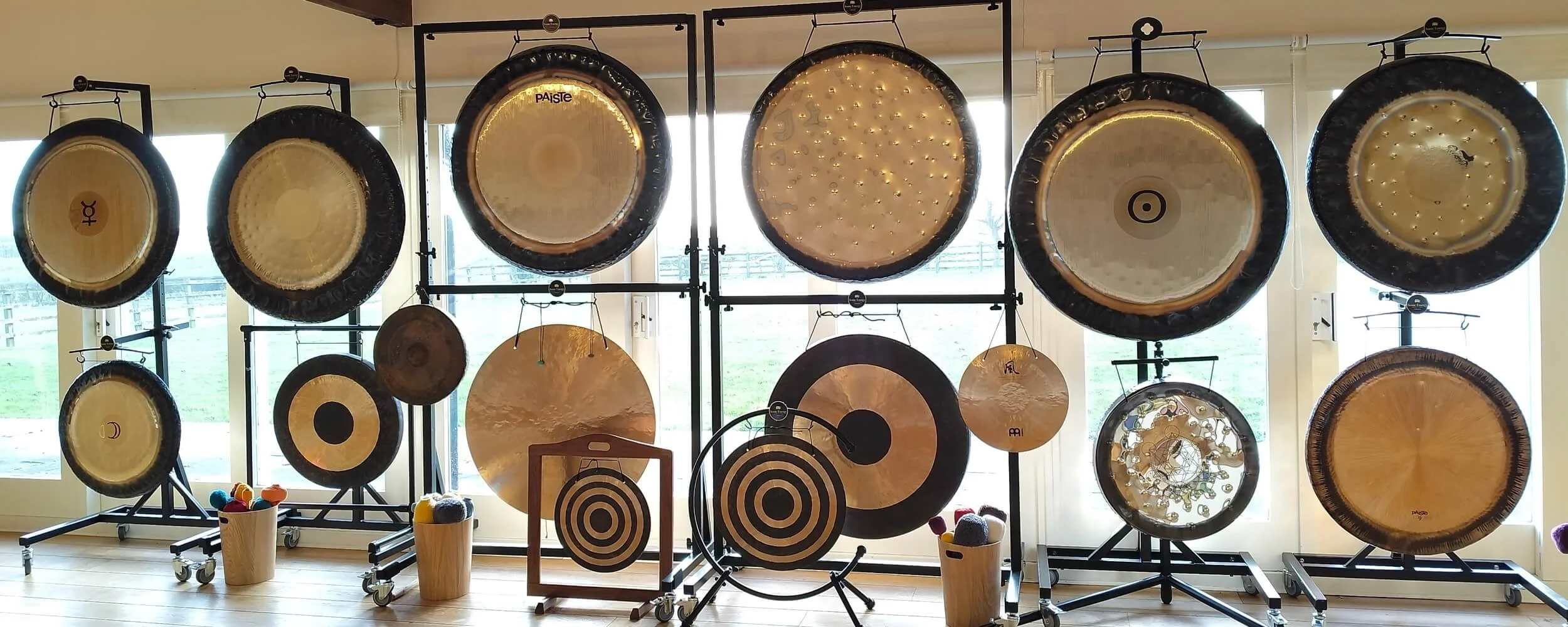 An extensive line of 16 professional master gongs arranged against the glass doors of our countryside training studio near Grantham, showcasing the specialized instrument selection available for advanced hands-on workshops.