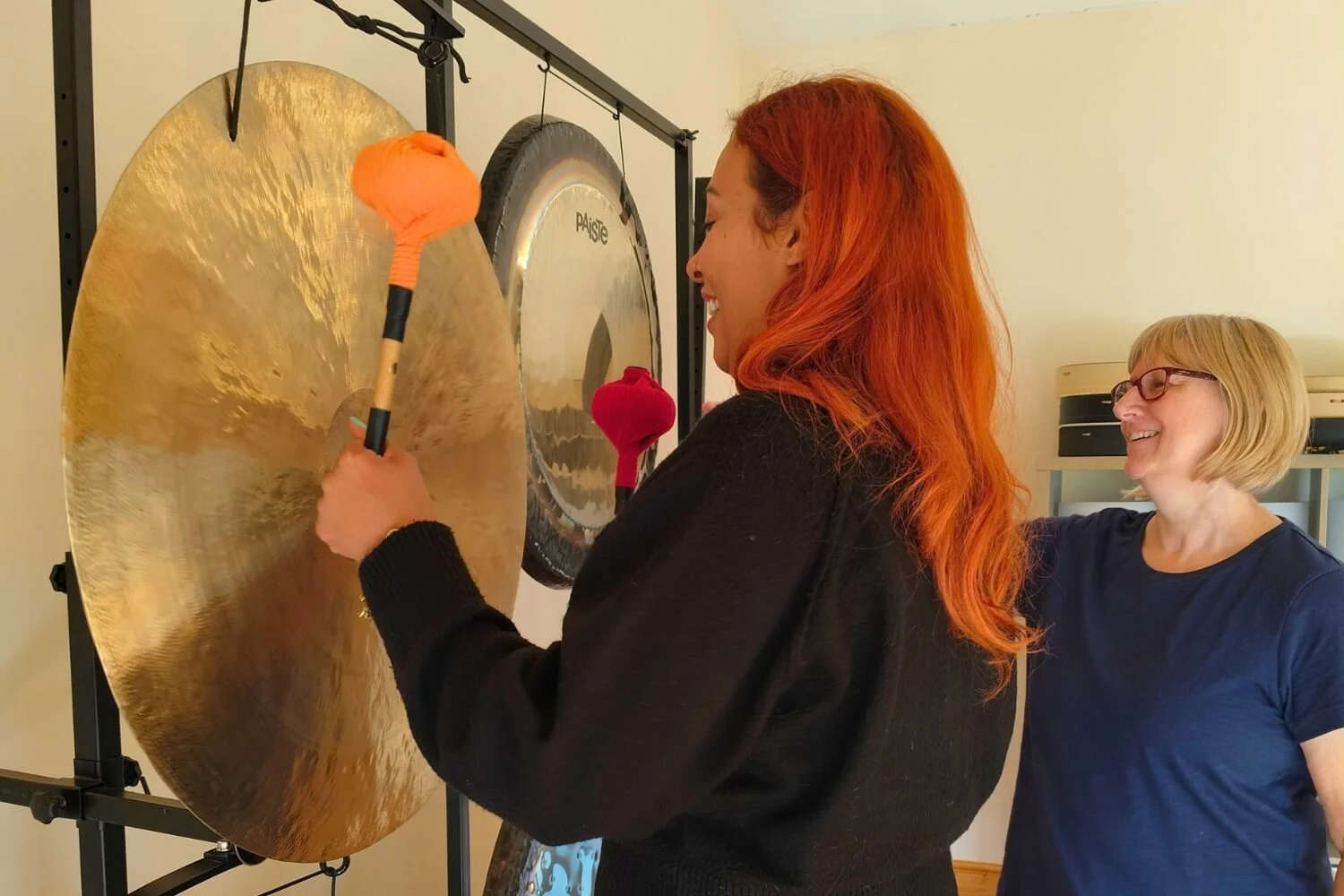 Lead tutor Helen Campbell smiling while teaching a student, Saffron, how to play a large wind gong using professional mallets against a neutral background.