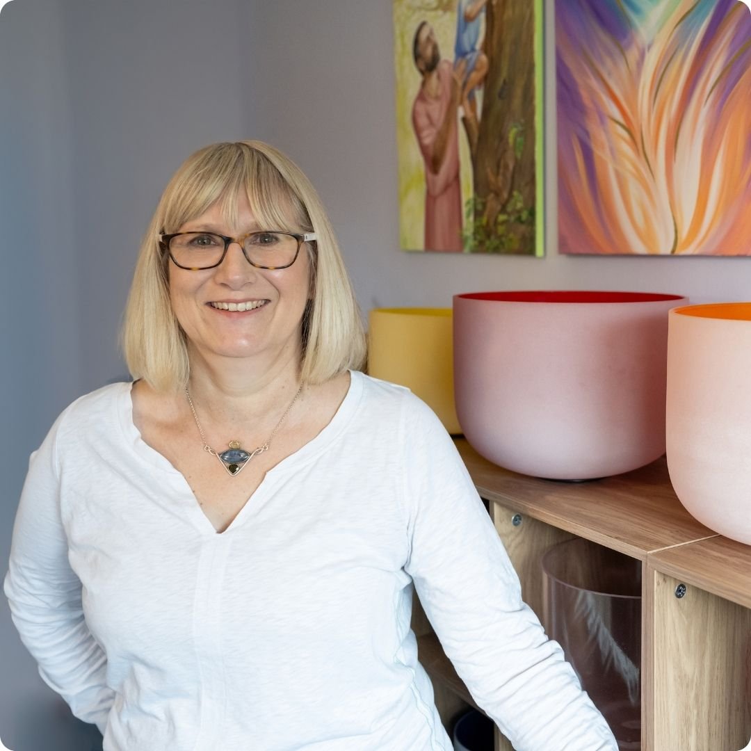 The tutor Helen. She is smiling with blonde hair and glasses and is standing in front of colorful artwork and crystal singing bowls on a wooden shelf.