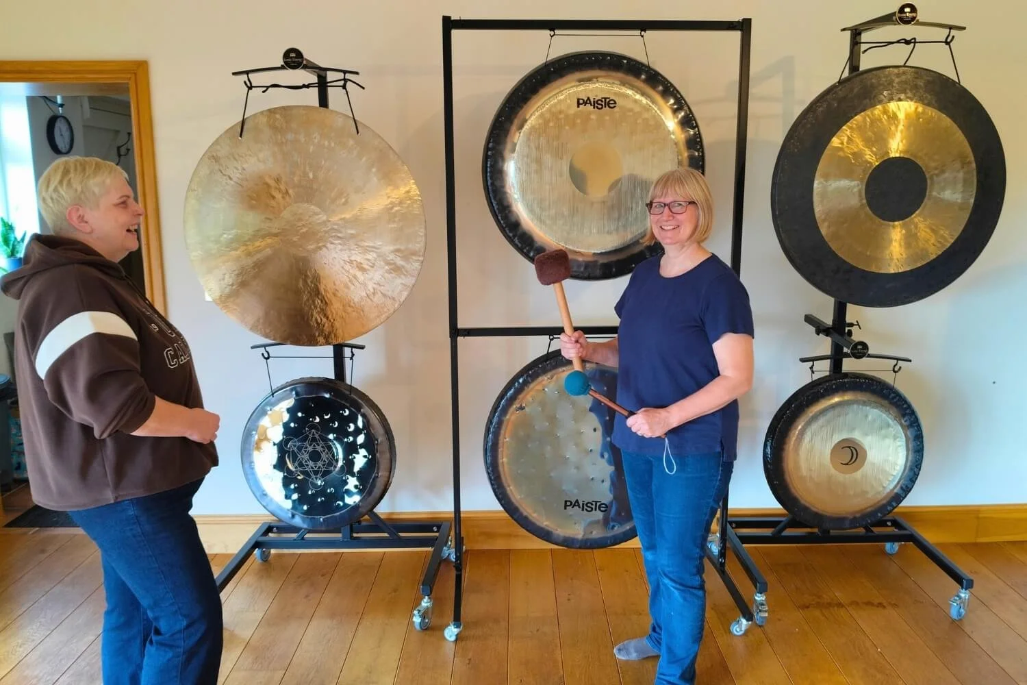 Sound therapy tutor Helen Campbell smiling and conversing with a student, Mandi, in front of a balanced alignment of six professional master gongs on a wooden floor.