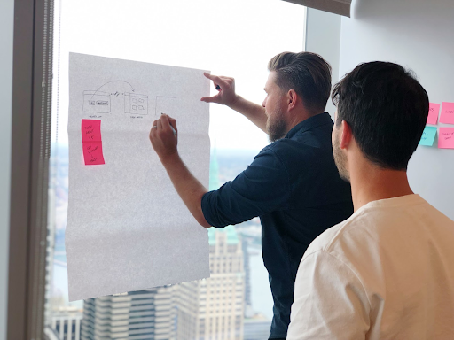 Two men discussing a large sheet of paper with sticky notes in a high-rise office overlooking a city.