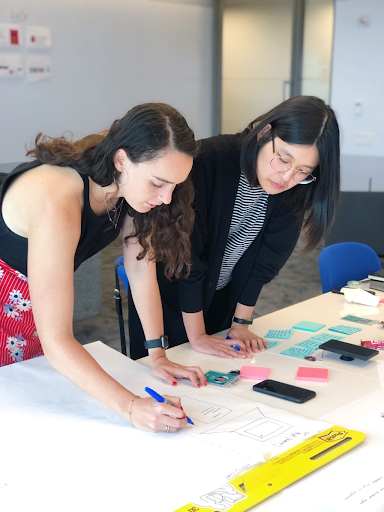 Two women working together on a design project at a table with papers, phones, and sticky notes in a modern office