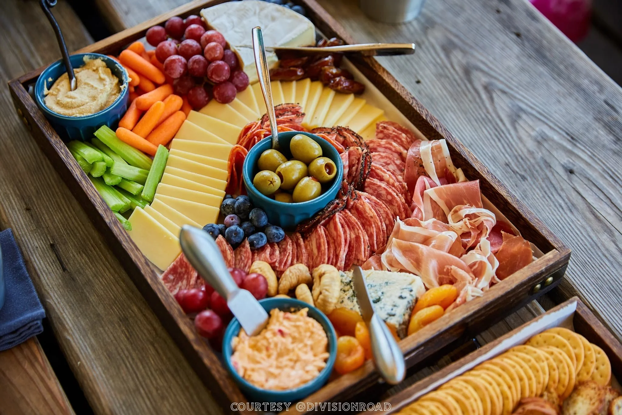 A charcuterie board with cheese, meats, grapes, blueberries, celery, carrots, olives, cracker chips, and dips on a wooden table.