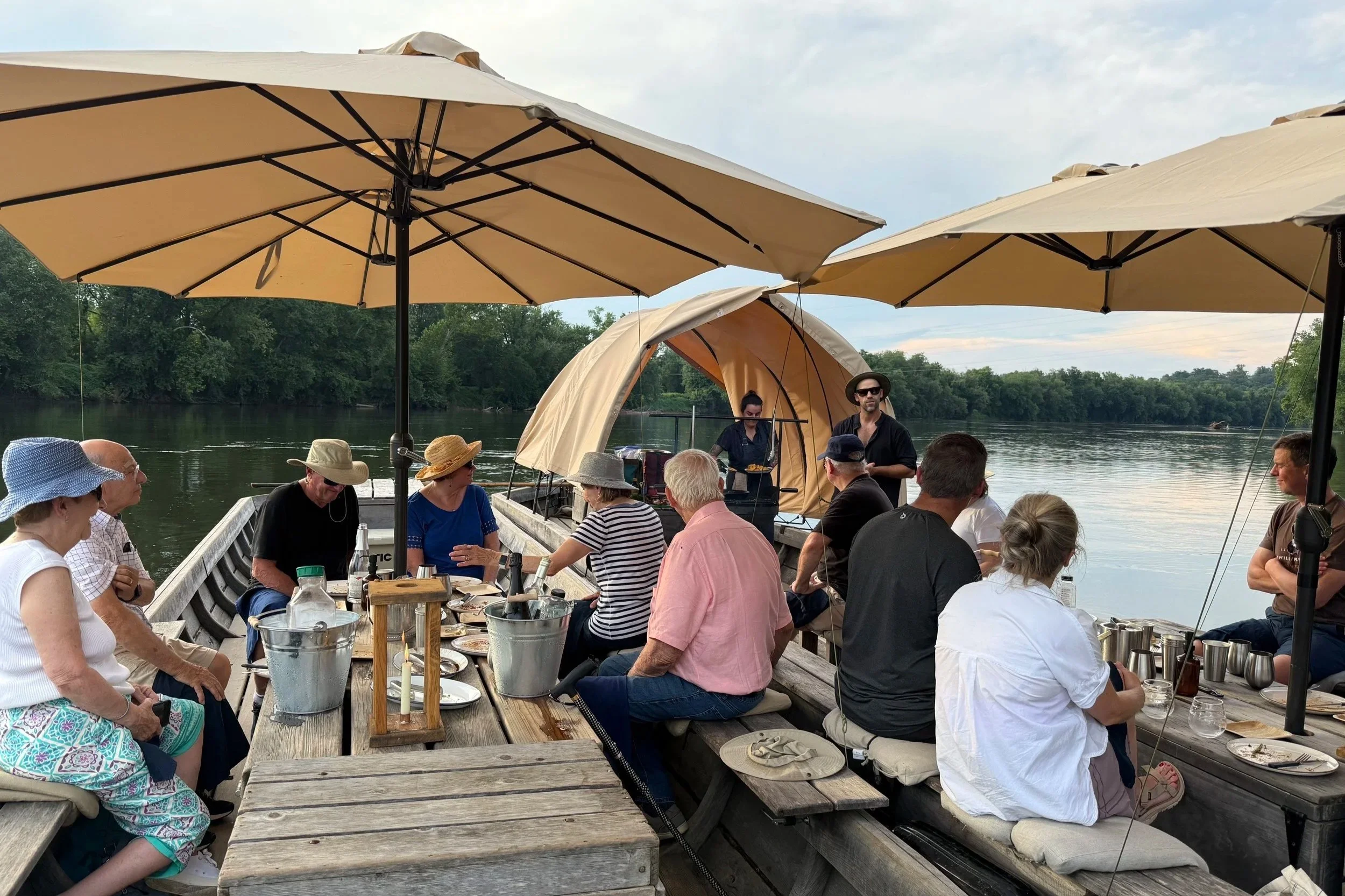 A group of people enjoying a gathering on a boat with a lakeside view, surrounded by trees, with large umbrellas providing shade and a small canopy tent set up on the boat.