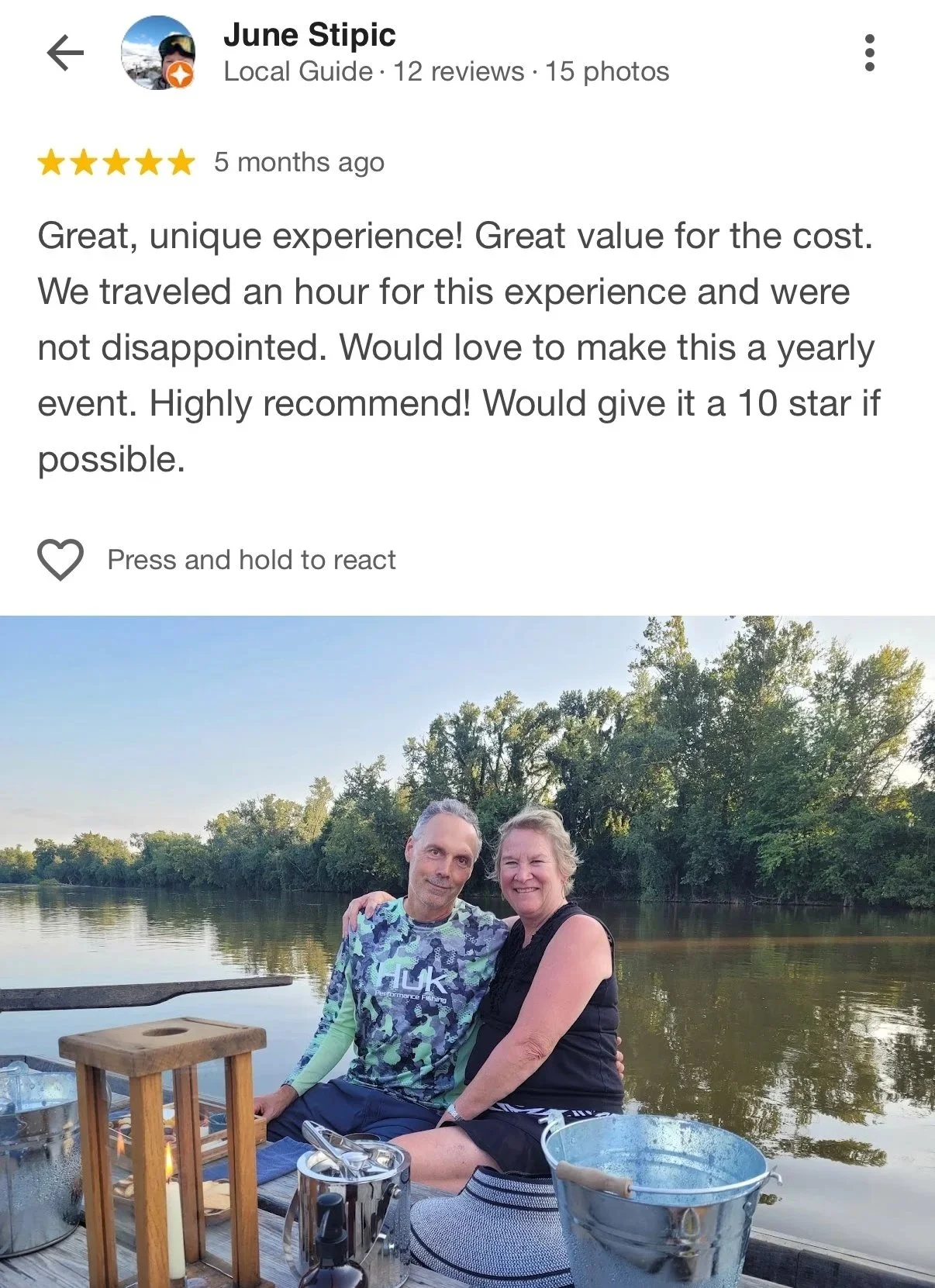 A photo of a man and woman sitting by a river at sunset, smiling at the camera. There are trees on the opposite bank, and they are on a wooden dock with a small table holding a lantern, drinks, and utensils.