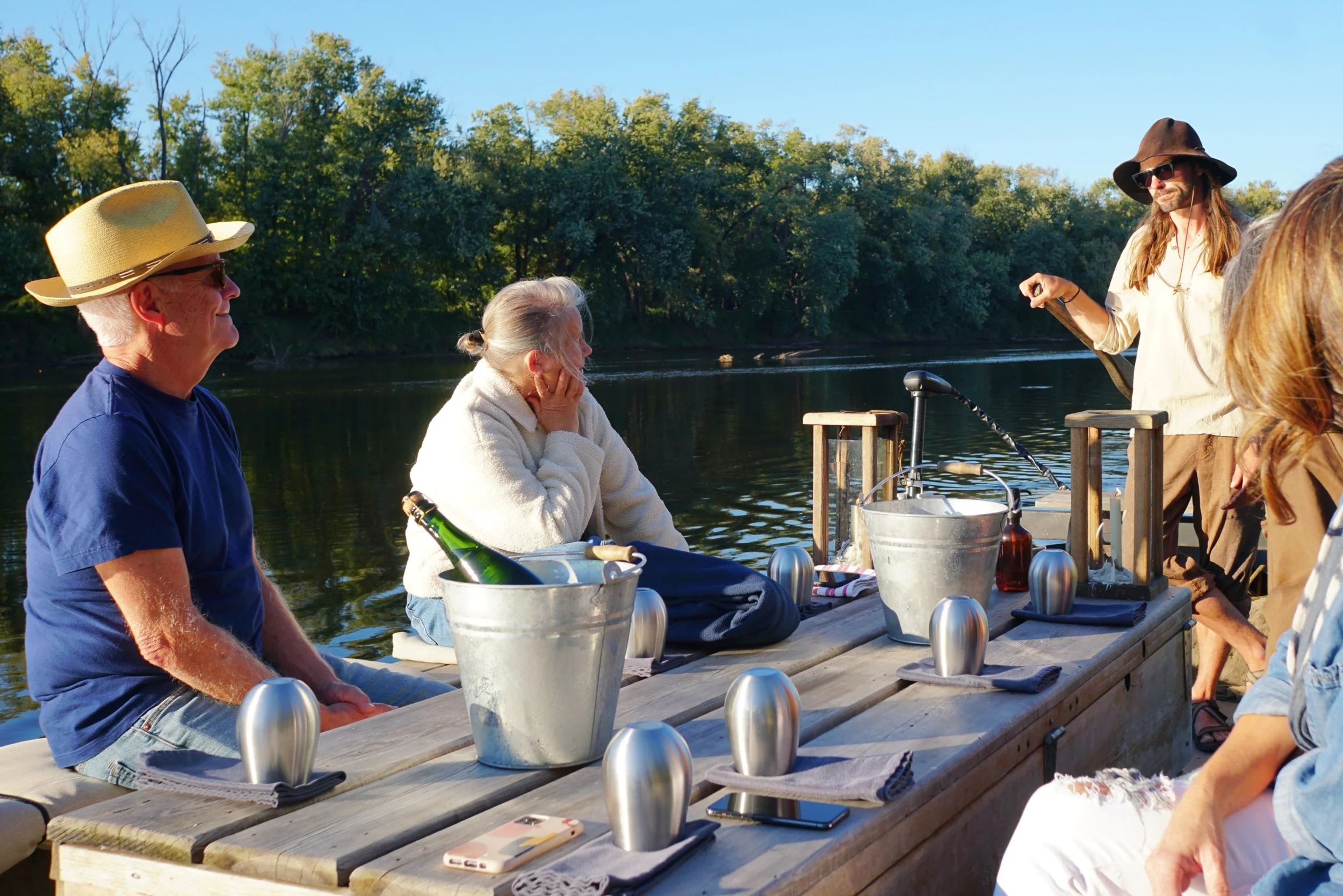 Group of people enjoying a boat trip on a lake surrounded by trees, with some sitting at a picnic table and others standing and chatting, sunlight casting warm glow.