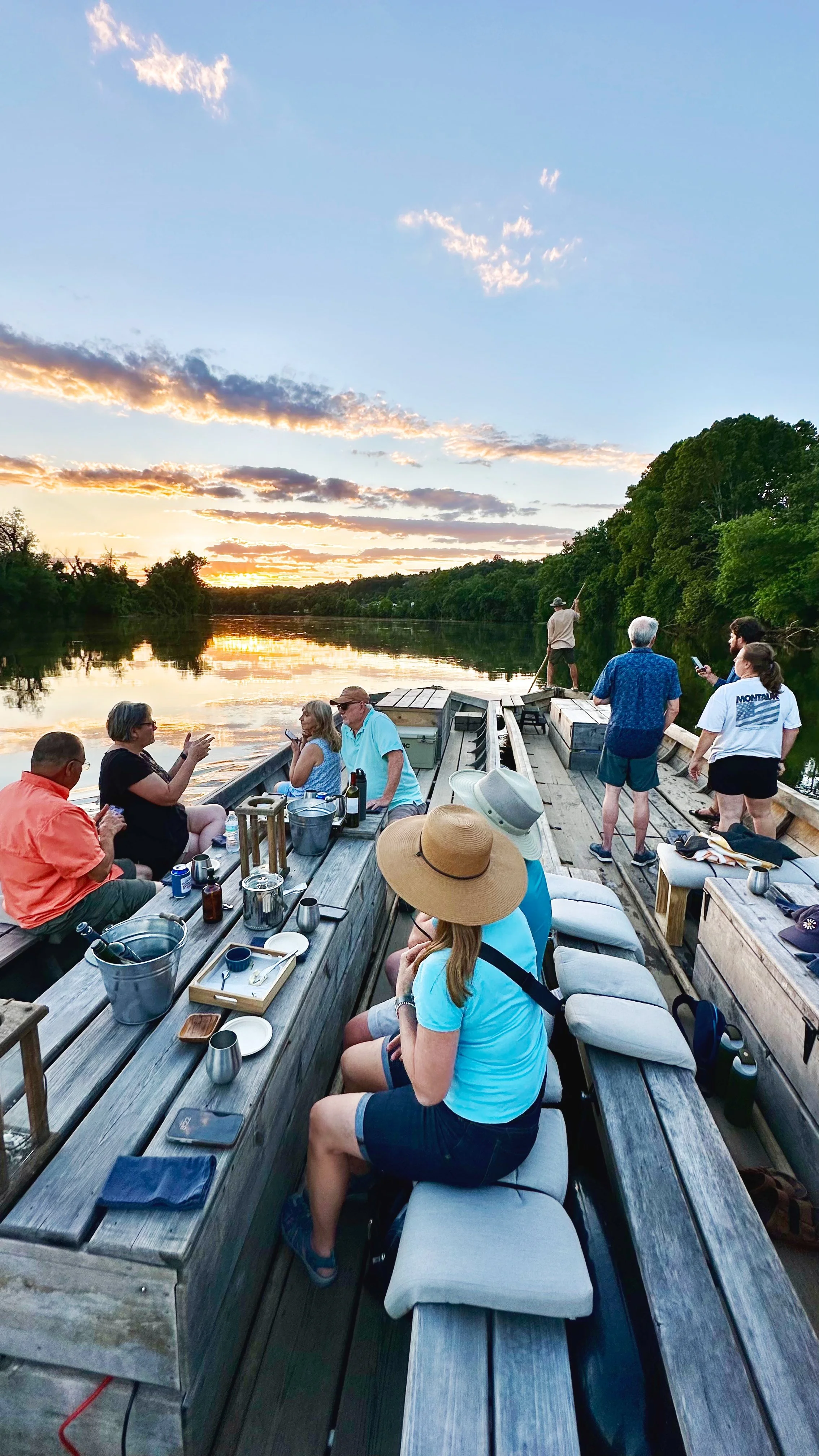 A group of people enjoying a social gathering on a boat during sunset on a calm river, with lush trees along the banks and a colorful sky with clouds.