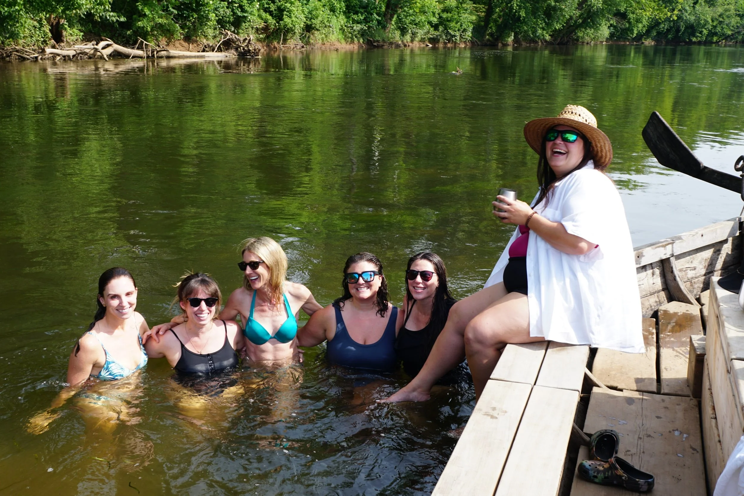 A group of six women enjoying themselves near a river. Five women are standing in the water, smiling, while one woman is sitting on the edge of a wooden boat wearing sunglasses, a sun hat, and a white cover-up, holding a tumbler.