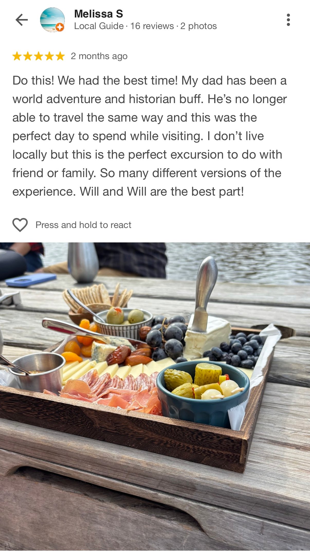 Charcuterie board with cheese, grapes, pickles, olives, meats, and crackers on a wooden outdoor table near a body of water.