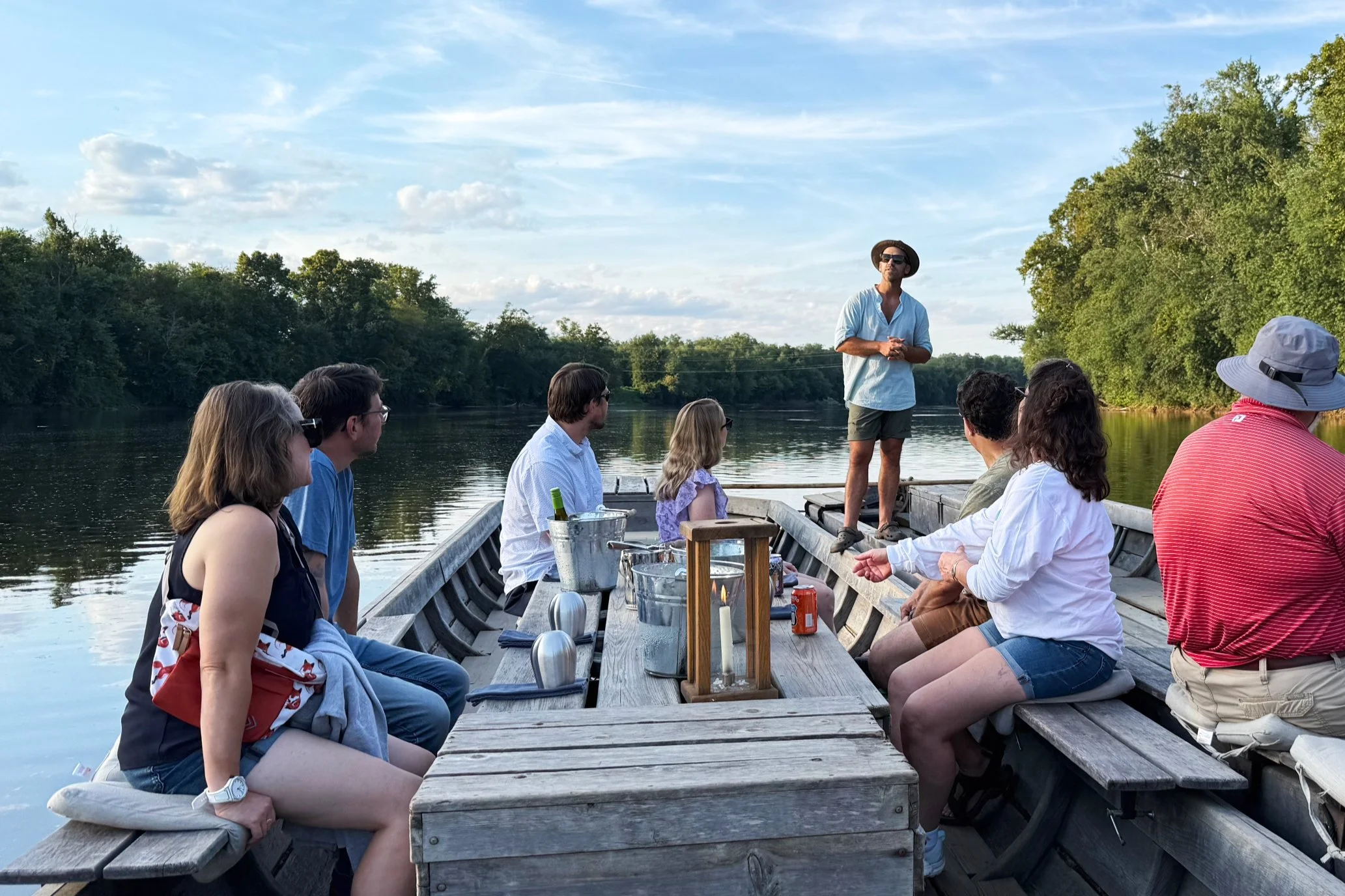 A group of people on a boat on a river, with a man standing and talking to them. The surrounding area is lush with trees, and the sky is partly cloudy.