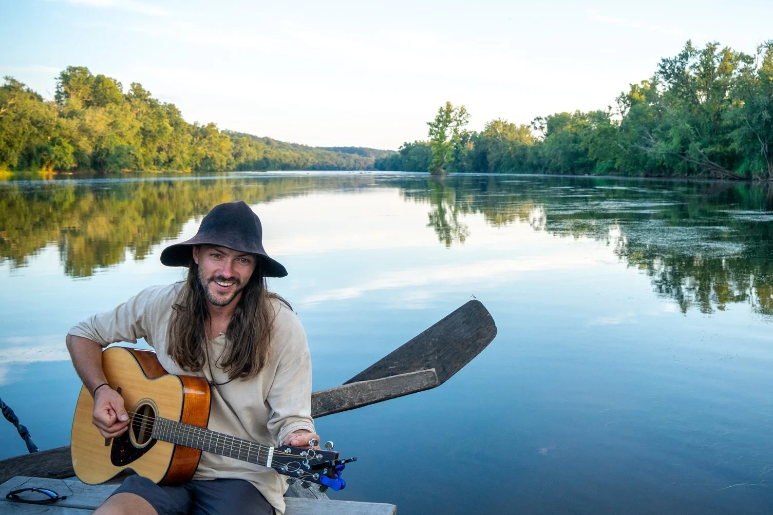 A man with long hair, beard, wearing a black wide-brimmed hat, beige long-sleeve shirt, and shorts, sitting on a wooden boat with a paddle, playing an acoustic guitar on a calm river with green trees on both banks under a clear sky.