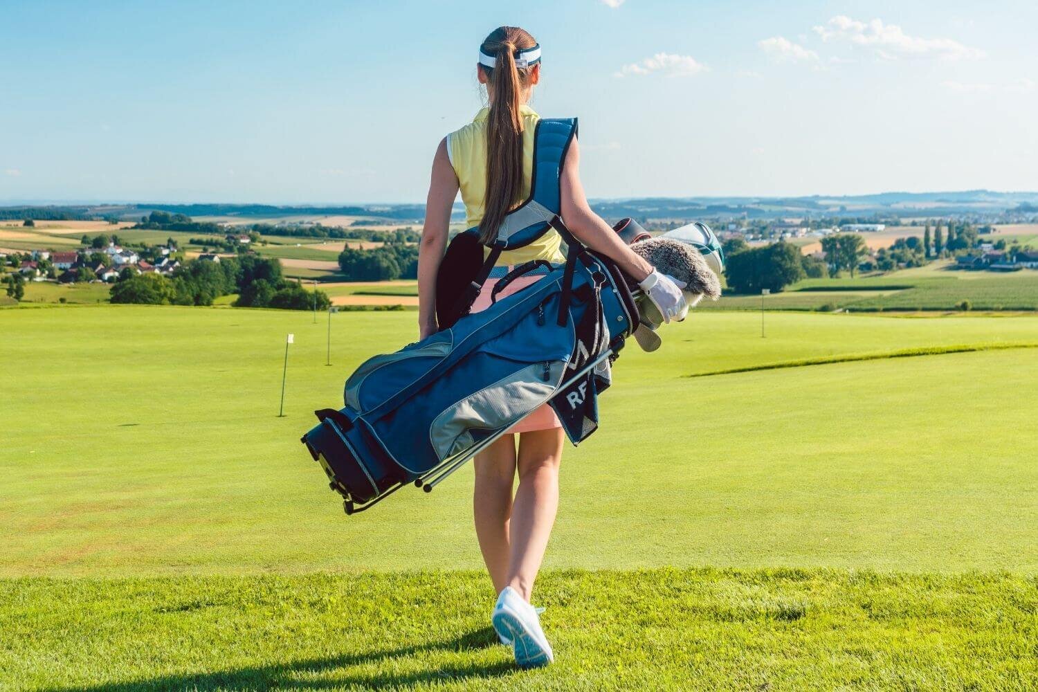 Woman golfer from behind dressed in golf attire carrying golf bag onto golf course