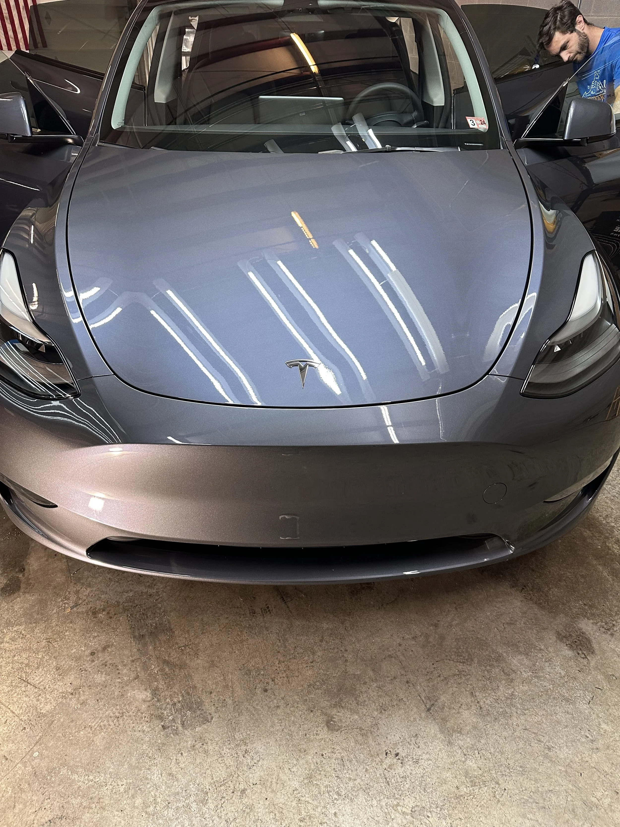 Front view of a dark gray Tesla electric car parked indoors, with a man in a blue shirt standing to the right, partially visible, inspecting or working on the vehicle.
