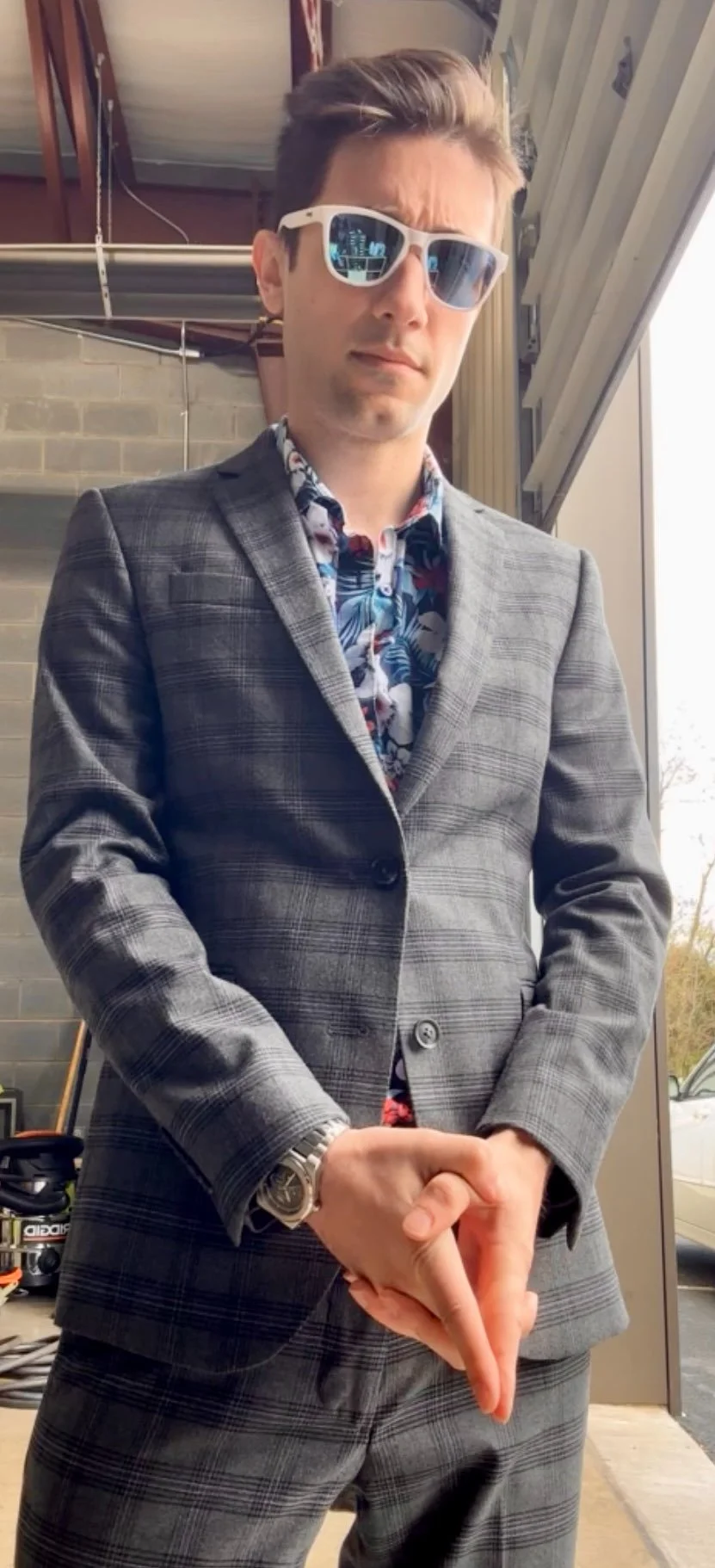 Young man wearing a gray plaid suit, floral shirt, white sunglasses, and a watch, standing indoors near a door with a brick wall in the background.