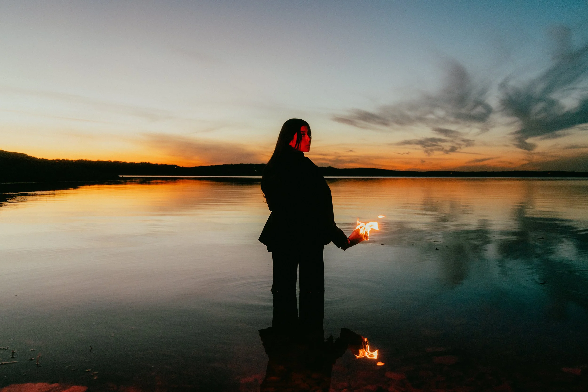 A person standing in shallow water at sunset, holding a small fire in their hand, with a silhouette against a colorful sky and its reflection in the water.