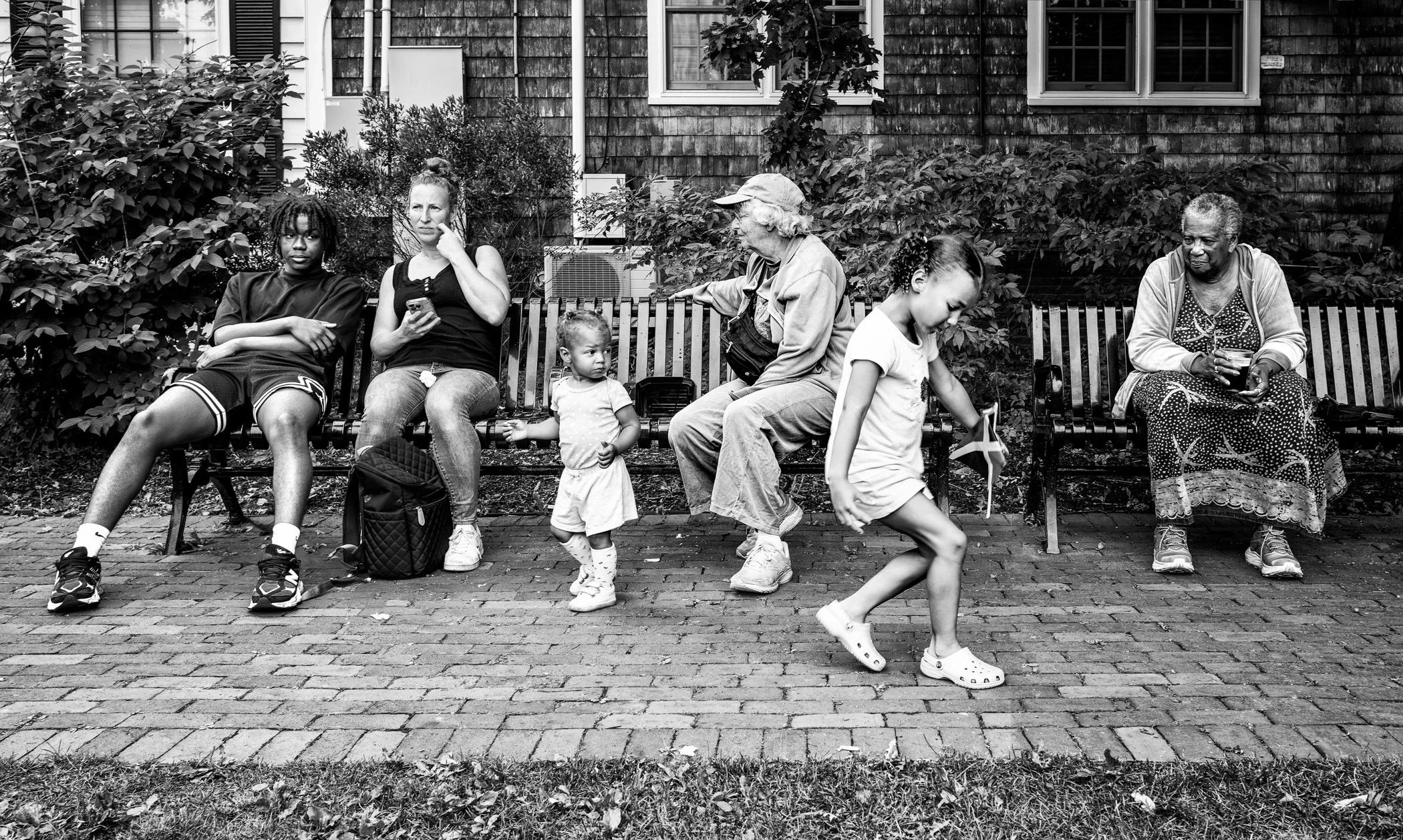 A park bench scene in black and white: There are two benches. On the first, there is a young Black man with twists, a white woman, a Black toddler, and an elderly white woman chatting. 2nd shows a Black woman smiling at a skipping Latin girl.