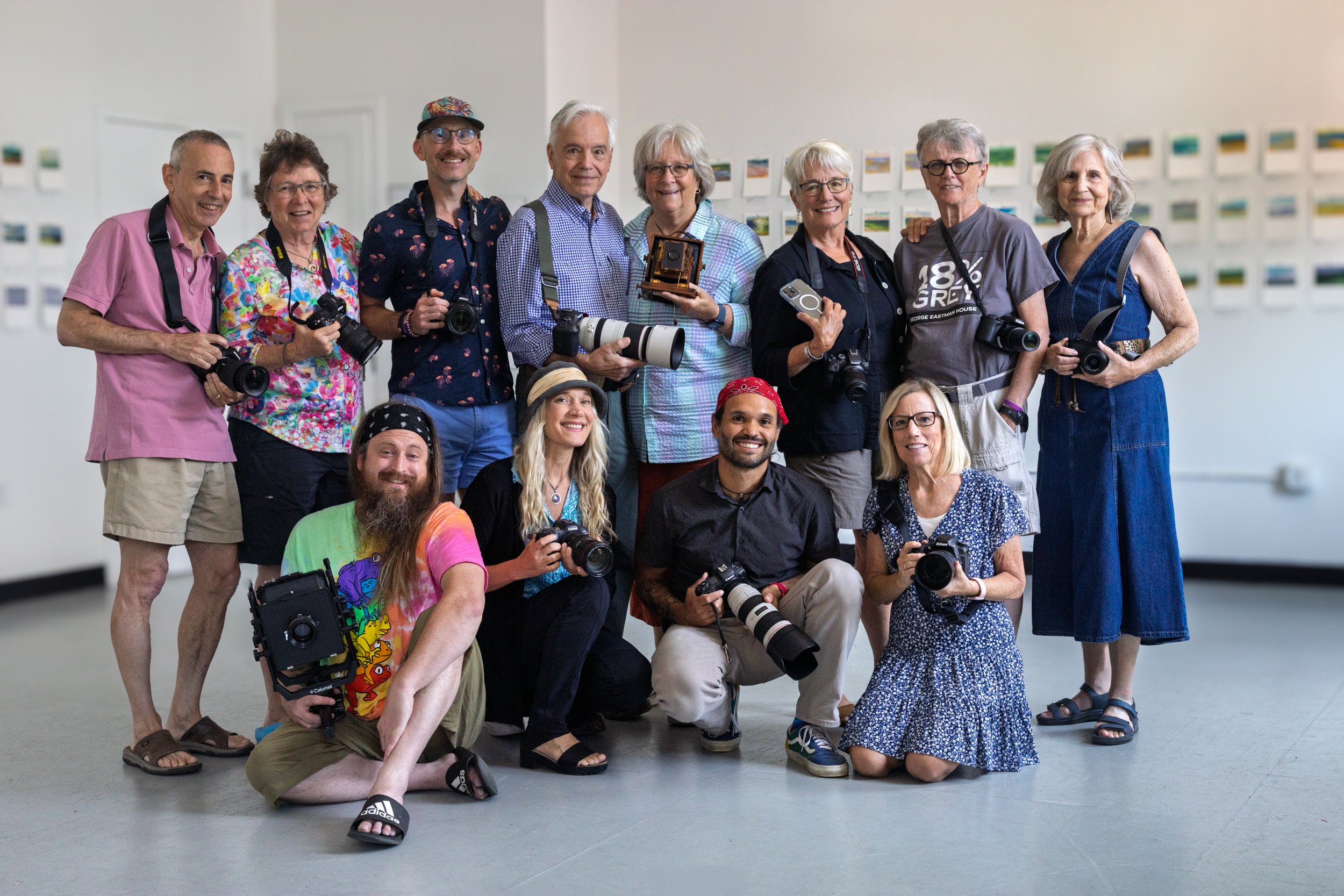 A group of 12 photographers are captured in a frame-lined gallery. They are all smiling and holding cameras. Four are seated or kneeling. Ten are standing behind.