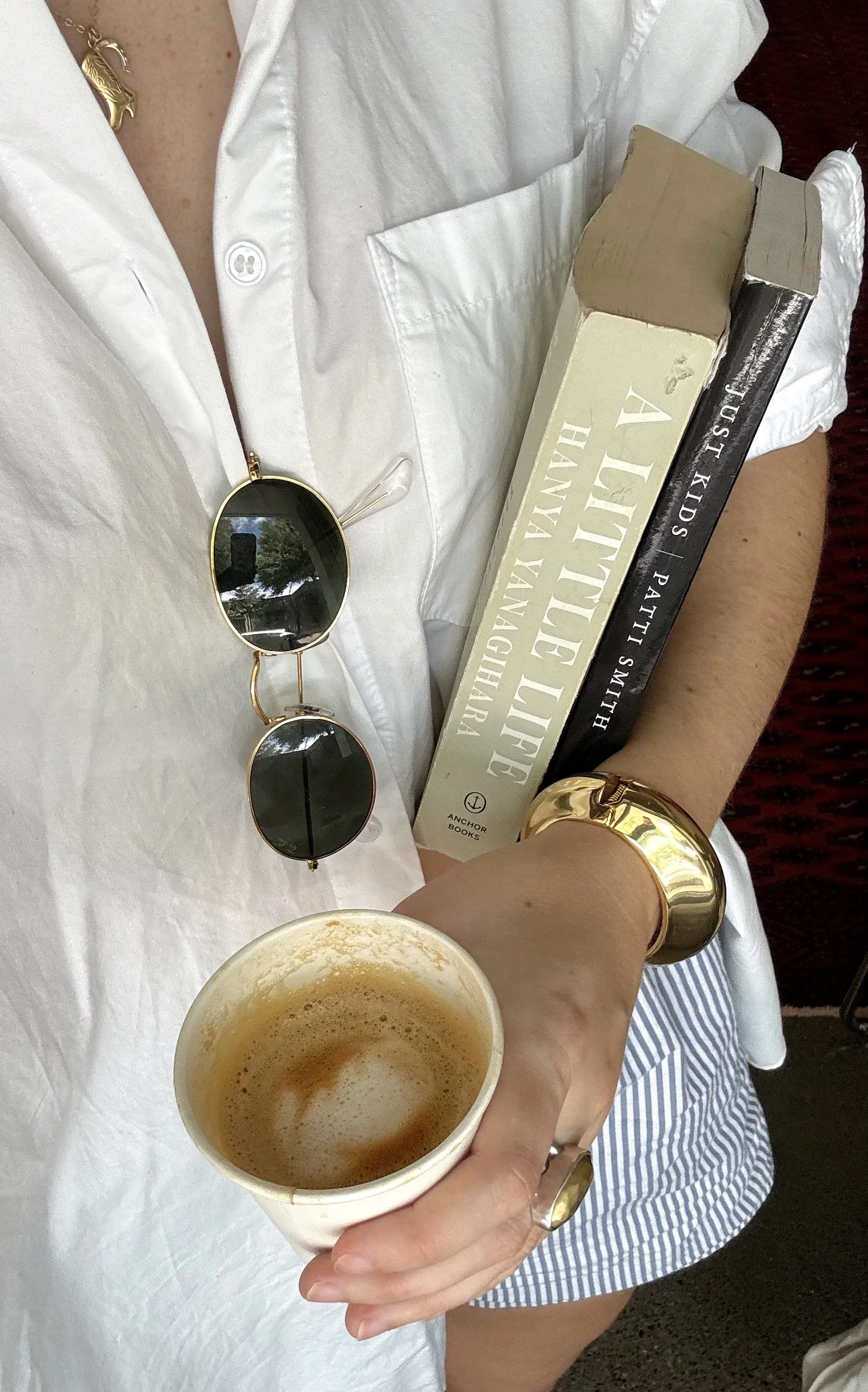 A woman wearing a white shirt, gold jewelry, and sunglasses holding a paper cup of coffee, with two books titled 'A Little Life' by Hanya Yanagihara and 'Just Kids' by Patti Smith resting on her arm.