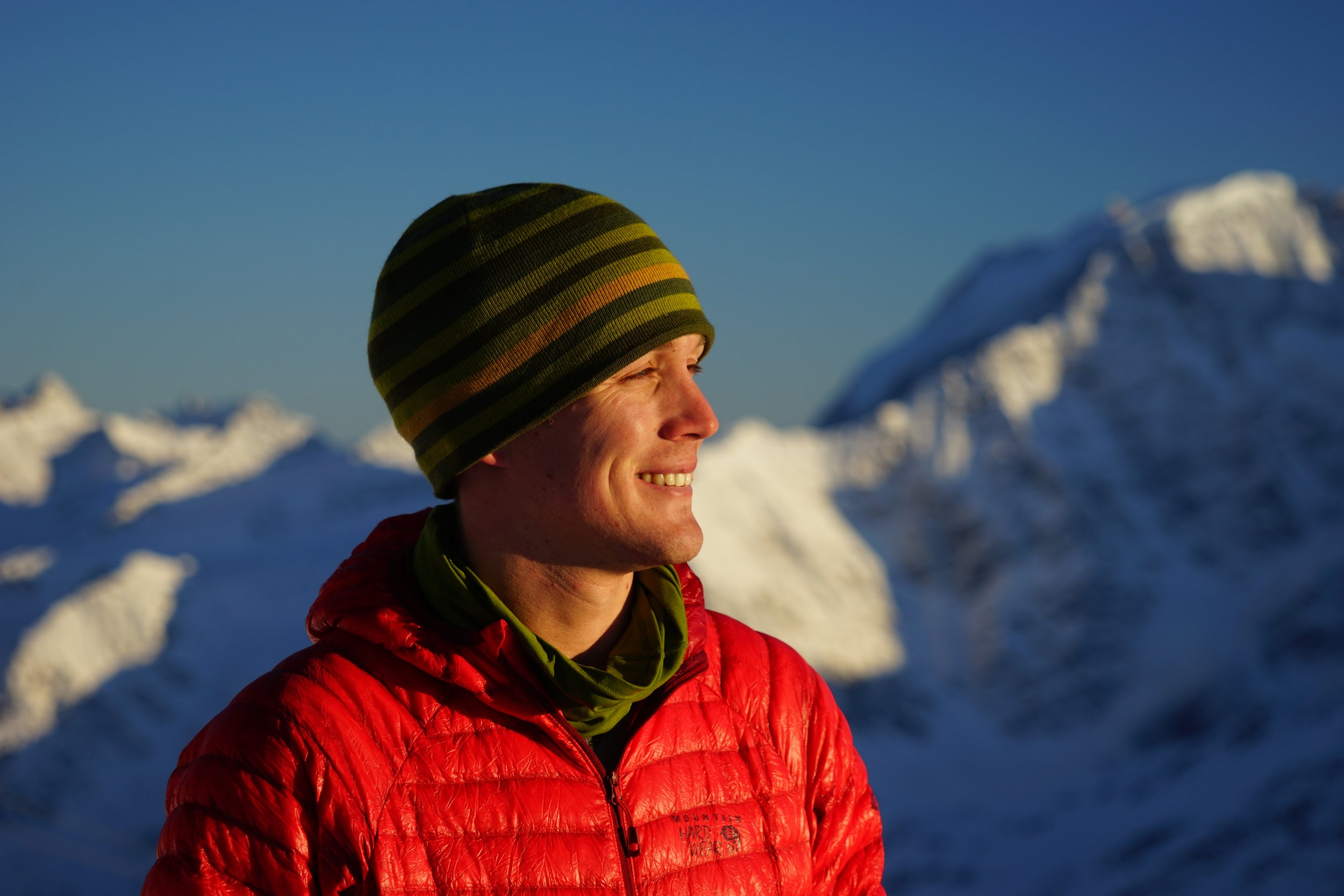 A man smiling outdoors wearing a striped beanie, bright red jacket, and green neck gaiter with snow-covered mountains in the background during the daytime.