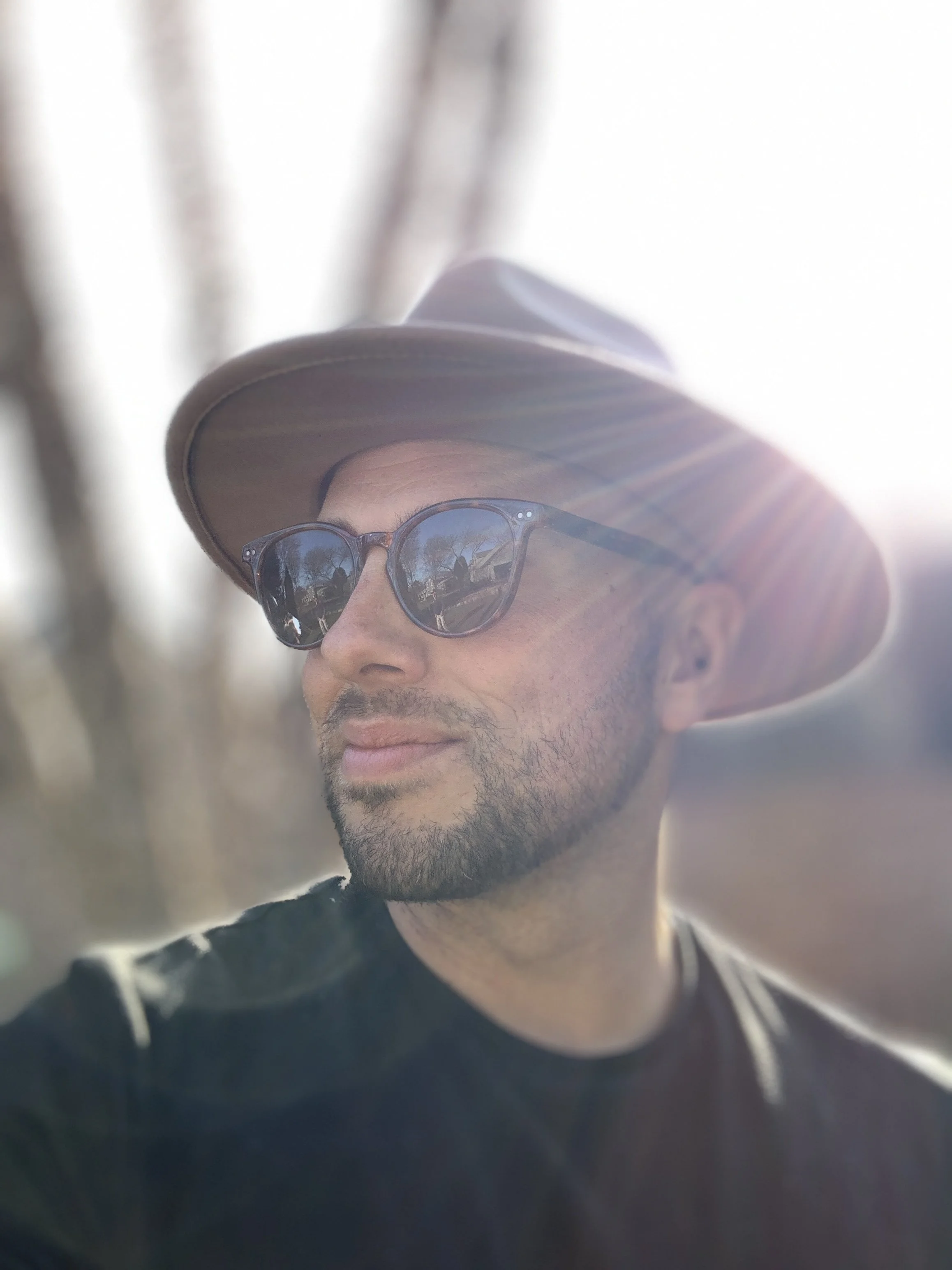 A man wearing sunglasses and a wide-brimmed hat outdoors, with sunlight creating a lens flare. He has a beard and is looking to the side with a gentle smile.