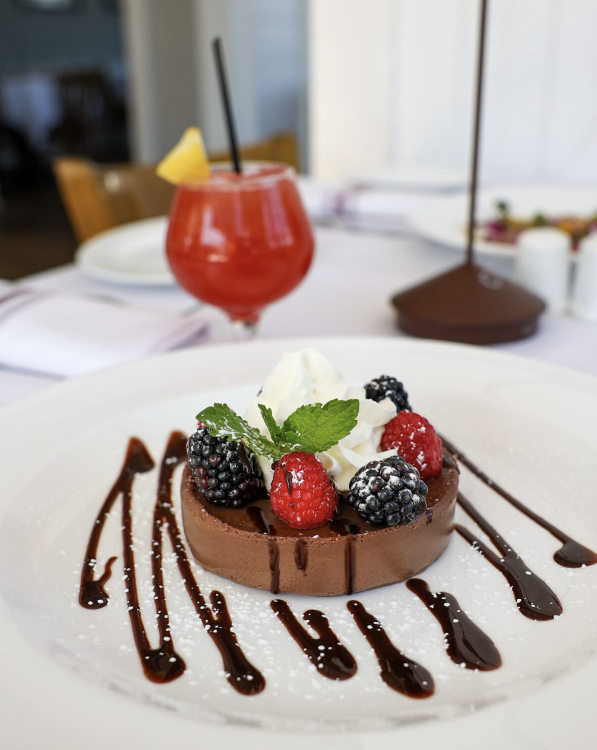 Chocolate tart topped with whipped cream, blackberries, raspberries, and mint, drizzled with chocolate sauce on a white plate.