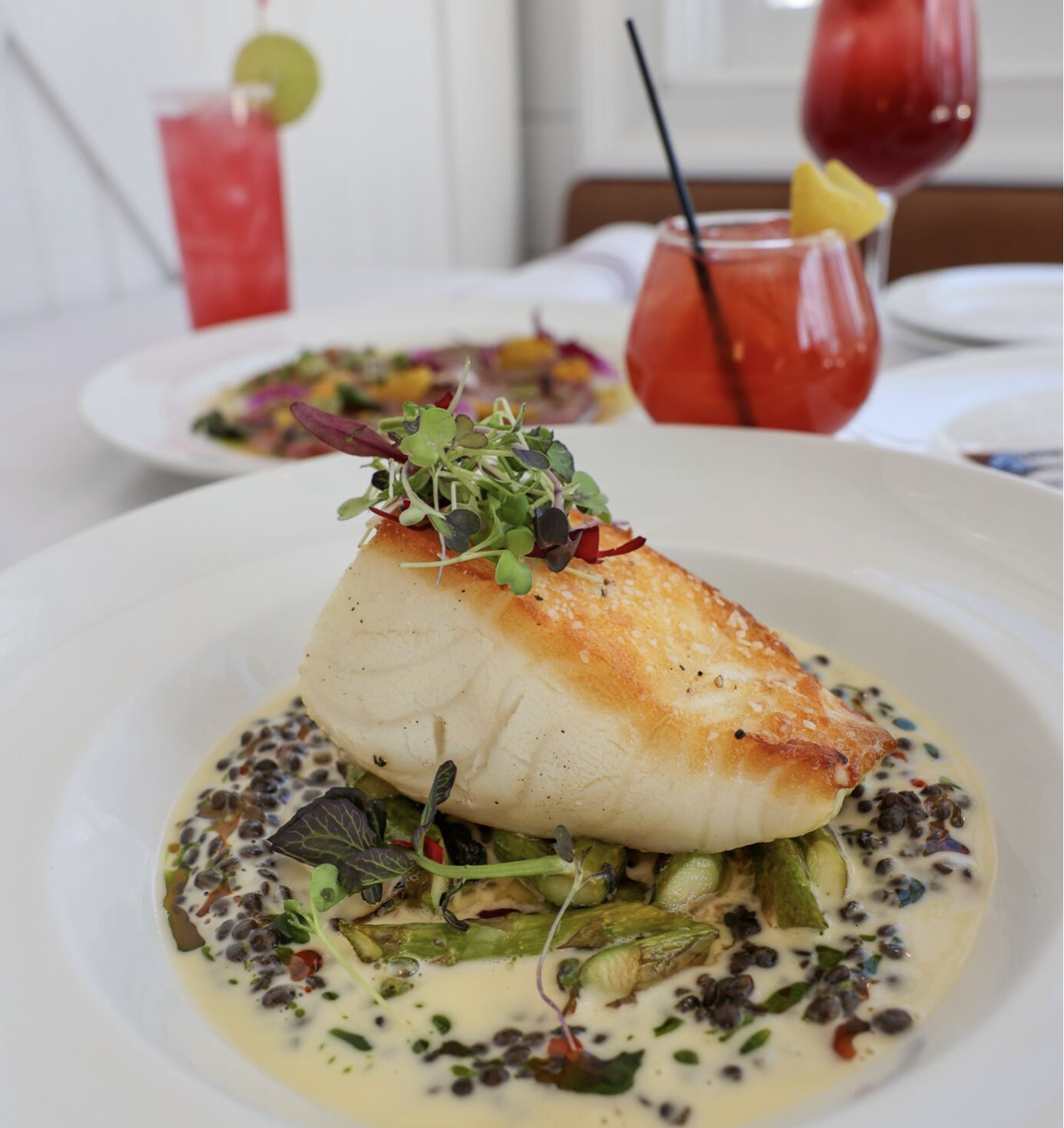 A plated dish of seared white fish on a bed of vegetables and creamy sauce, garnished with microgreens, with two beverages in the background on a dining table.