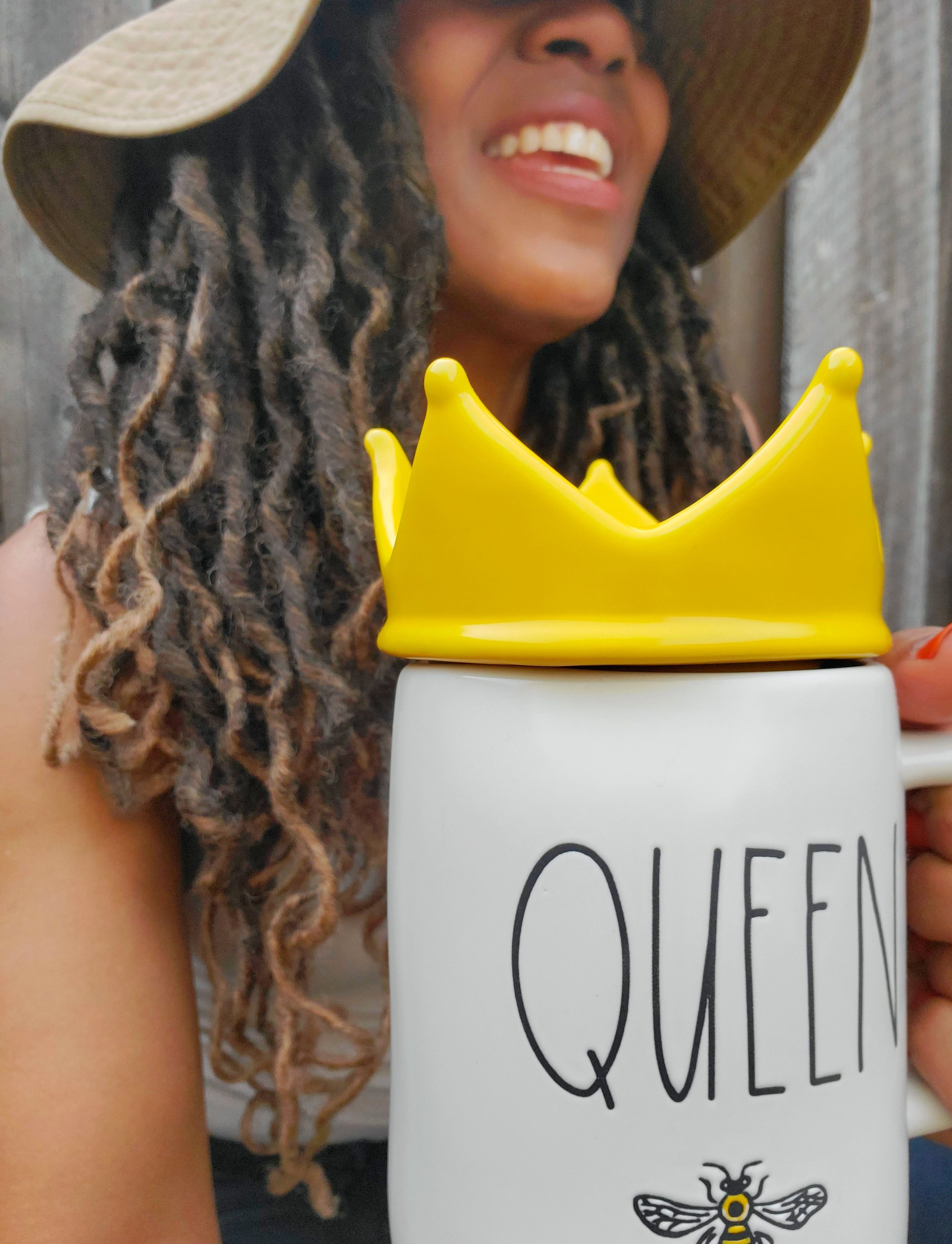 Person wearing a hat holding a white mug with the word 'QUEEN' and a bee graphic, featuring a yellow crown lid.